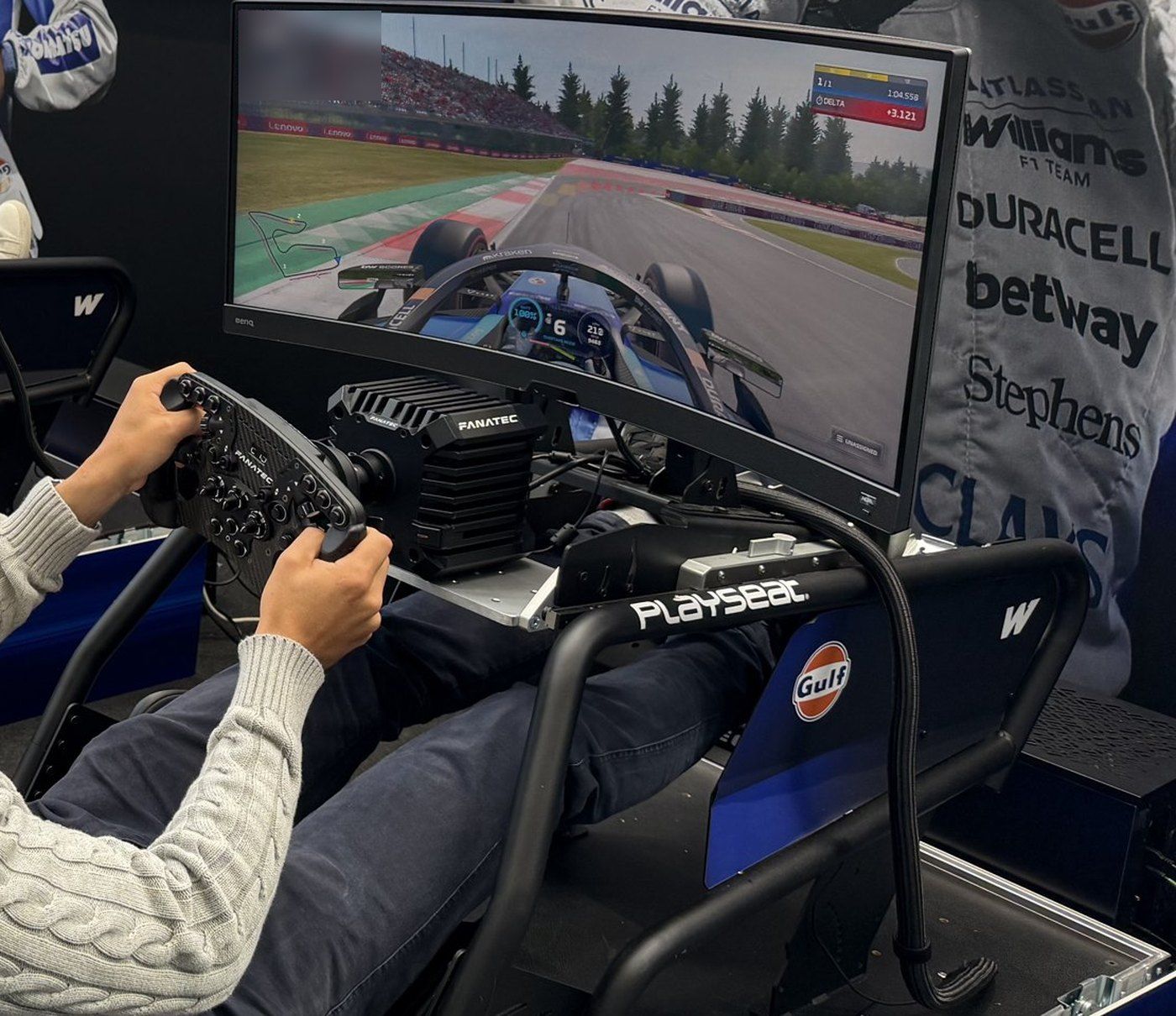 A sim racing rig seen over the driver's shoulder: hands on a racing wheel, a curved screen showing an F1 game in a blue car, with Williams sponsor branding on the wall behind