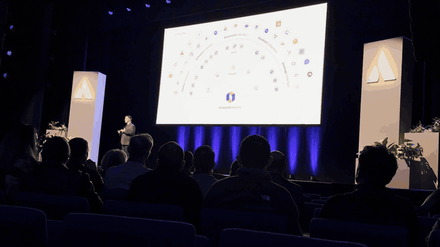 A dark auditorium seen from among the audience at Partner Accelerate: a speaker on stage in front of a large bright slide with app icons arranged in arcs, and the audience in silhouette
