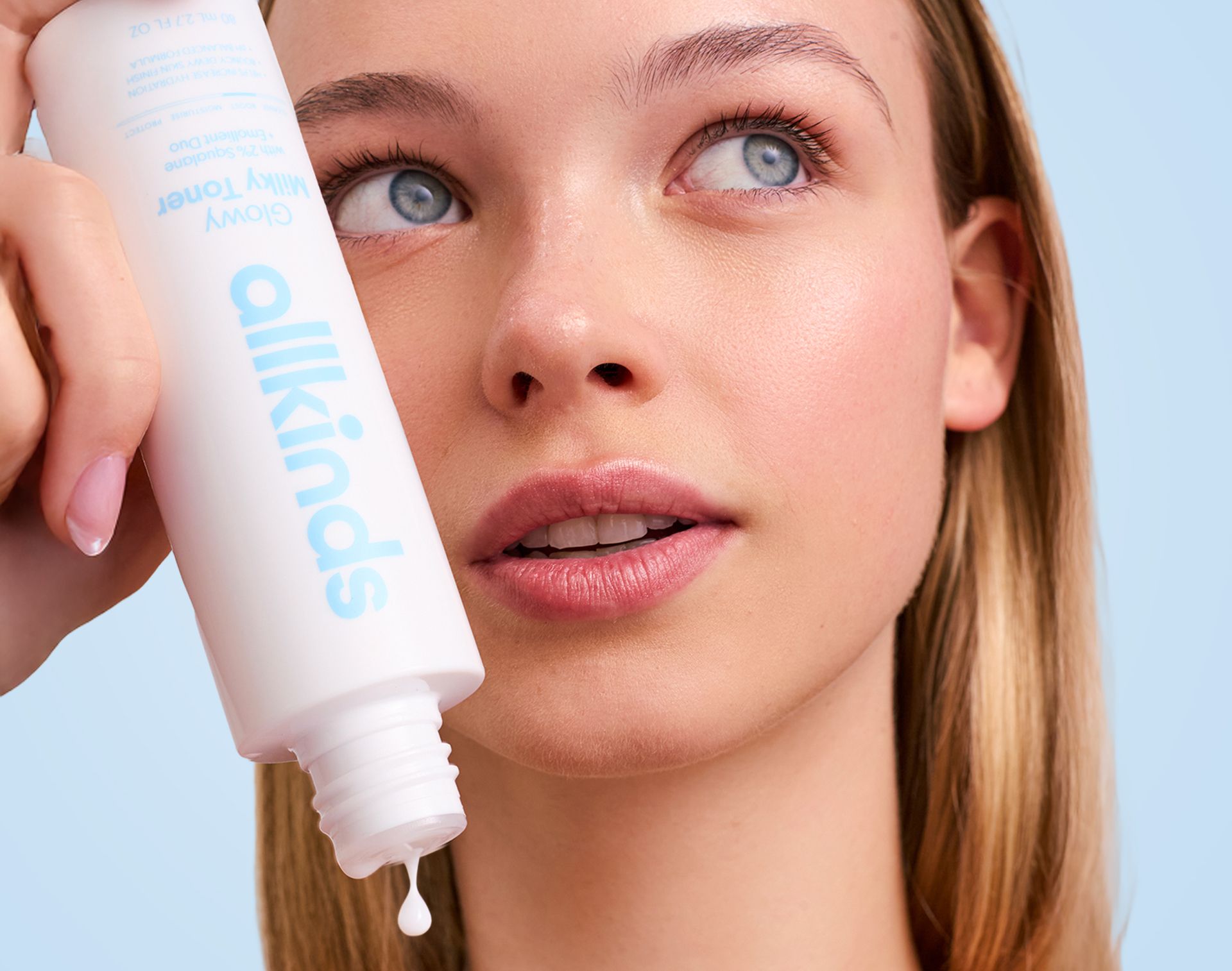 Woman holding an allkinds Glowy Milky Toner bottle with a drop dripping, looking up.