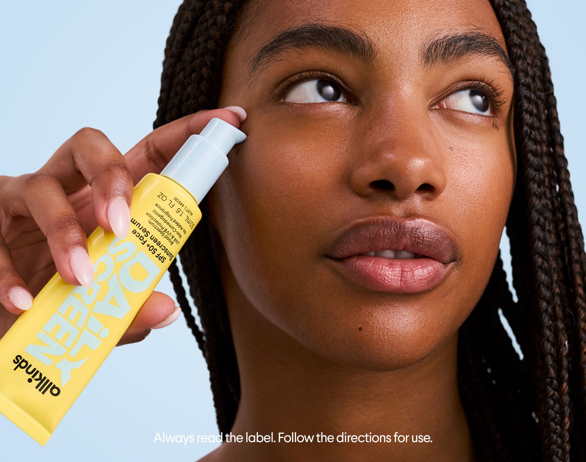 A person with dark skin applies Allkinds SPF 50+ Face Sunscreen Serum from a yellow tube near their temple.