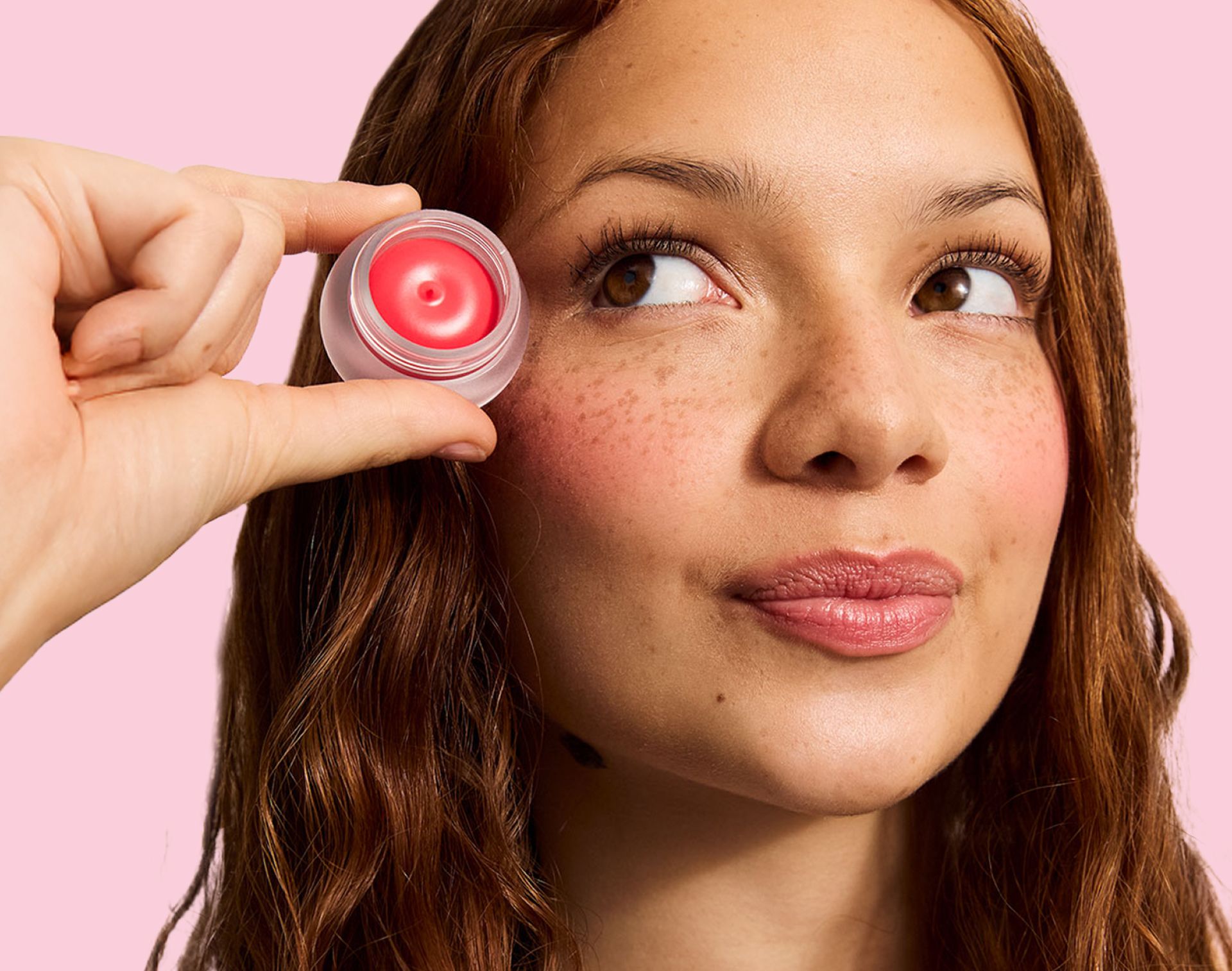 A woman with freckles and brown hair looks up while a hand holds a small pot of coral pink balm near her temple.
