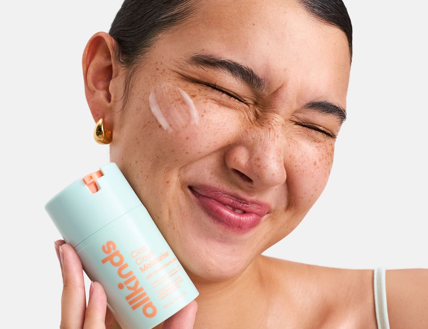 A woman with freckles playfully squeezes her eyes shut, holding an "allkinds" moisturizer bottle with product on her cheek.