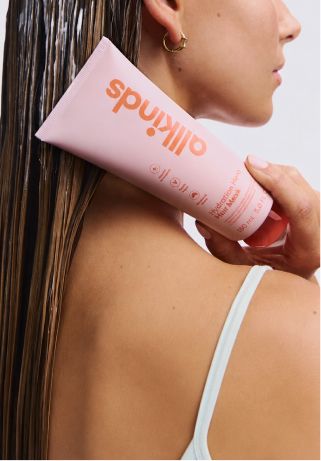 A person with wet hair holds a pink tube of allkinds Hydration Hero Hair Mask.