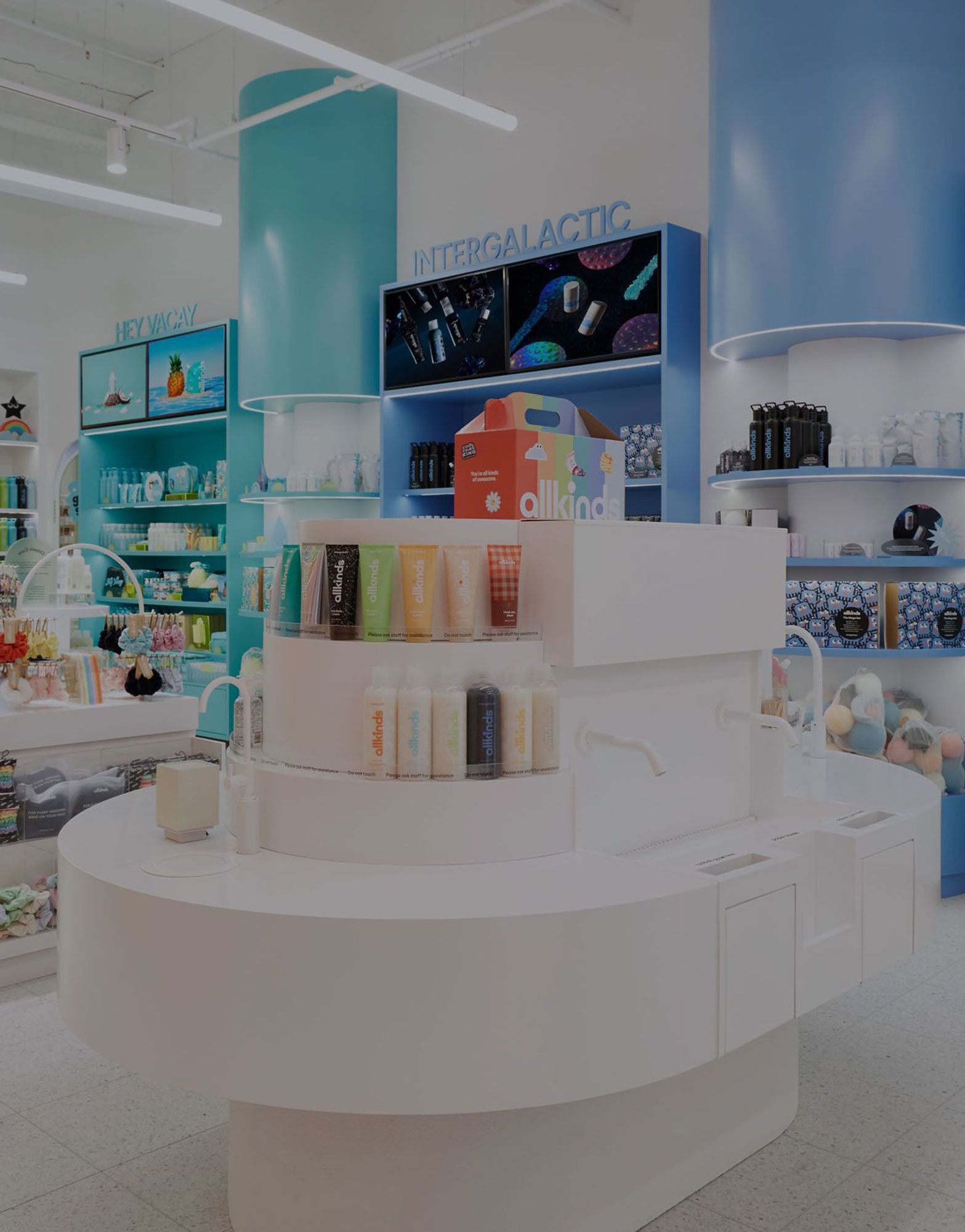 Bright, modern store interior with a white tiered display featuring allkinds products, blue and teal accents, and digital screens showing "HEY VACAY" and "INTERGALACTIC" content.