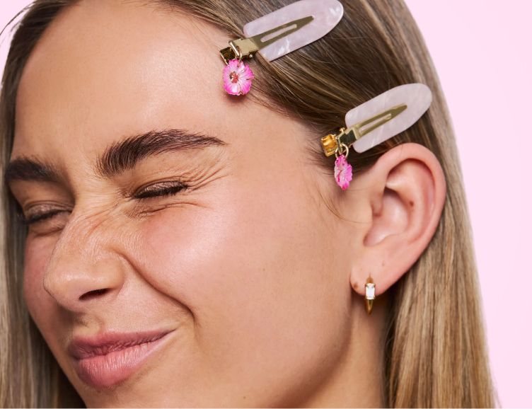 A woman squints her eyes shut, wearing two pink flowered hair clips and a small gold earring.