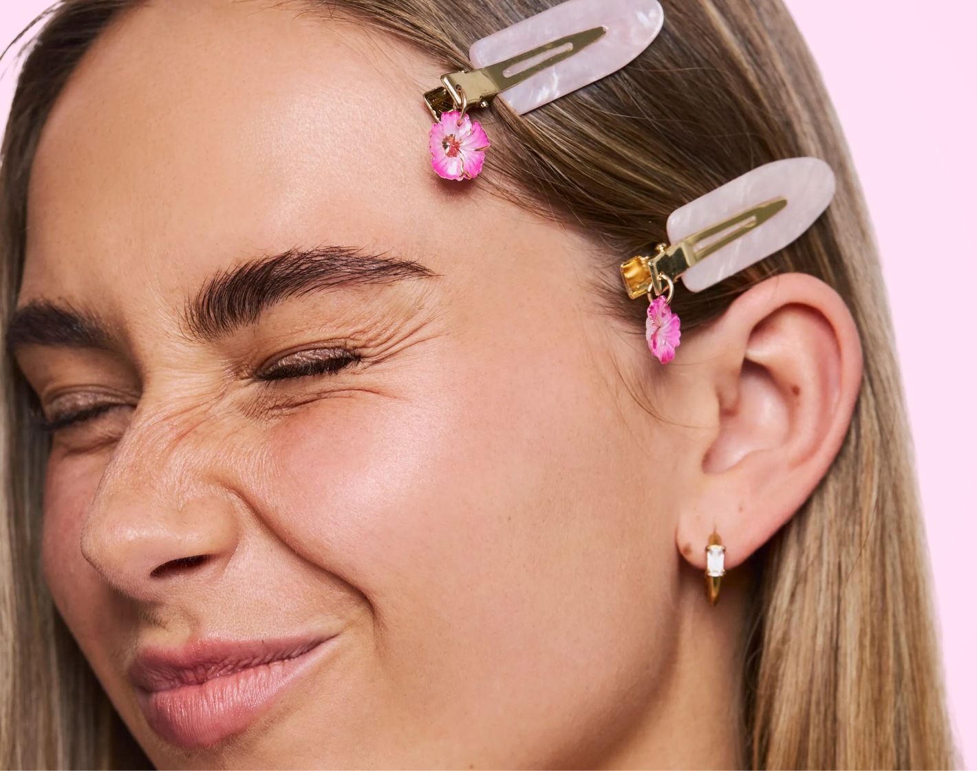 Close-up of a woman with a playful squint, wearing two hair clips with pink flower charms and a gold earring.