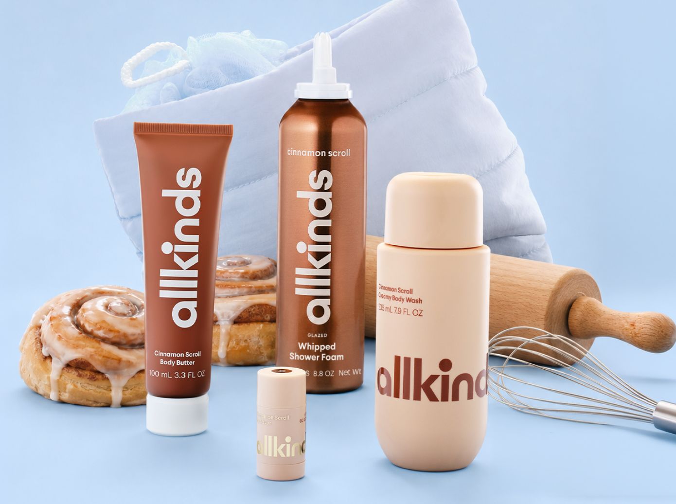 Allkinds cinnamon scroll scented body care products (body butter, shower foam, body wash, and stick) displayed with real cinnamon rolls, a rolling pin, whisk, and towel on a blue background.