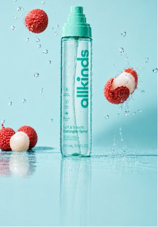 A clear "allkinds Soft & Smooth Detangler Spray" bottle with teal accents, surrounded by lychees and water splashes on a light blue background.