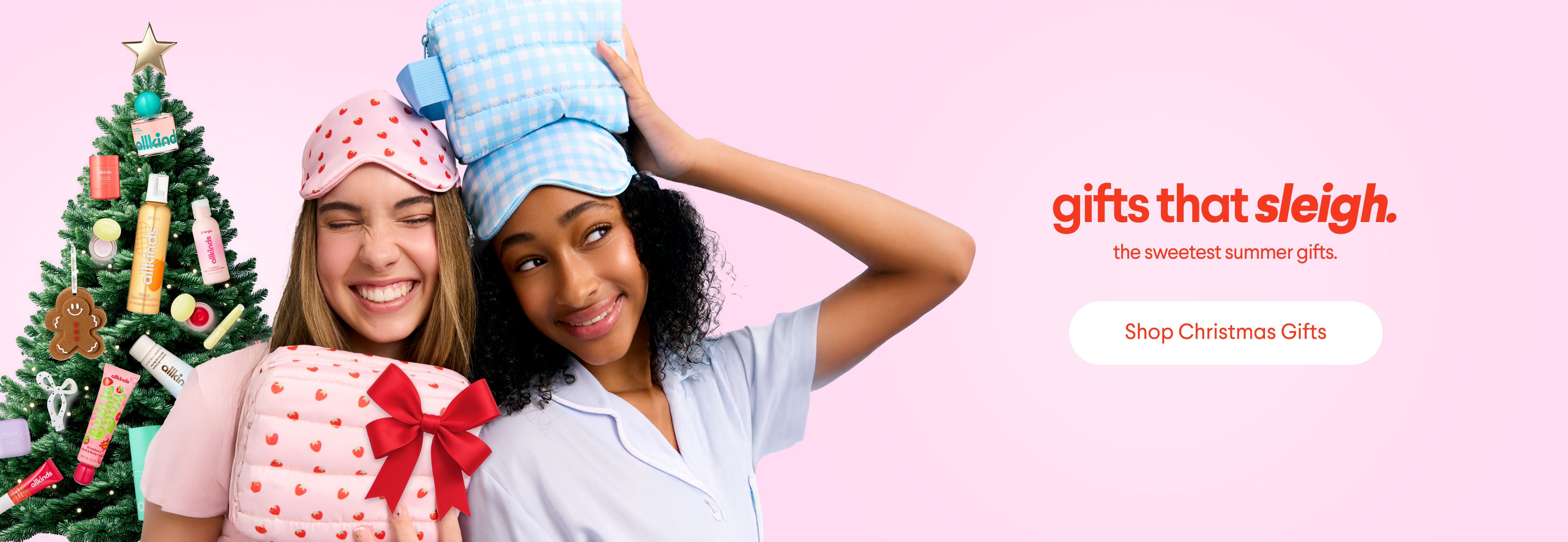 Two smiling young women in pajamas and sleep masks, one holding a gift, stand next to a Christmas tree decorated with beauty products. Text says "gifts that sleigh. the sweetest summer gifts." with a "Shop Christmas Gifts" button.