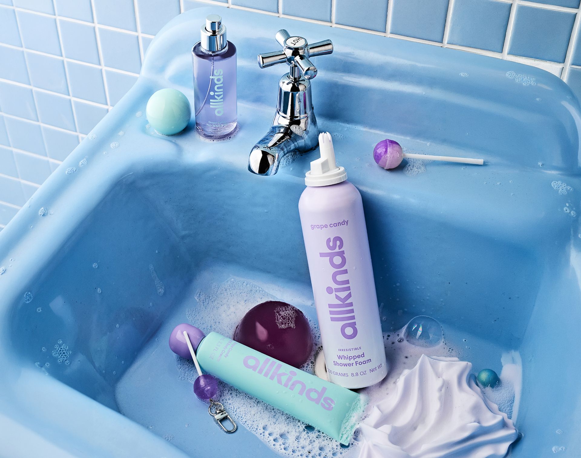 Allkinds bath and body products, bath bombs, and lollipops fill a bubble-filled blue double sink.