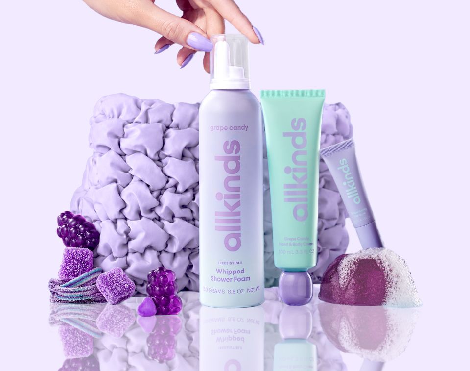A hand reaches for Allkinds lavender whipped shower foam, mint green hand cream, and purple lip gloss, displayed with grape candies, a fizzing purple bath bomb, and ruffled lavender fabric on a reflective lavender surface.