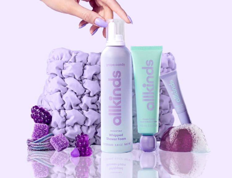 A hand holds a purple Allkinds shower foam, a mint green cream stands next to purple candies and a fizzing bath bomb on a reflective surface.