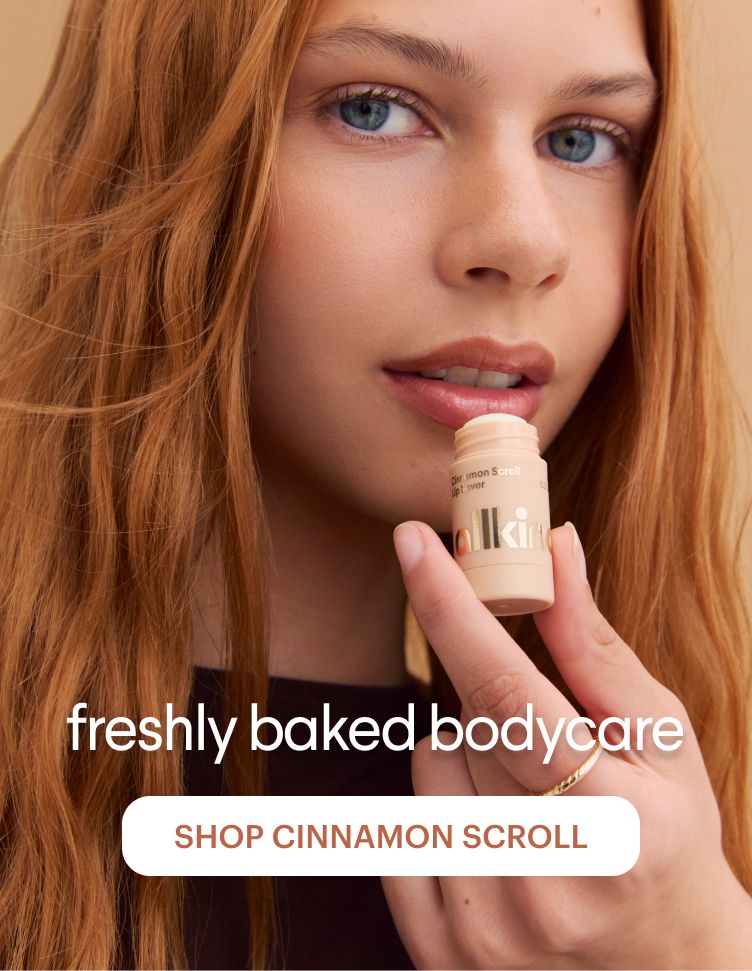 A woman with long red hair holds a "Cinnamon Scroll Lip Lover" stick near her lips. Overlaid text reads "freshly baked bodycare" and a button says "SHOP CINNAMON SCROLL".