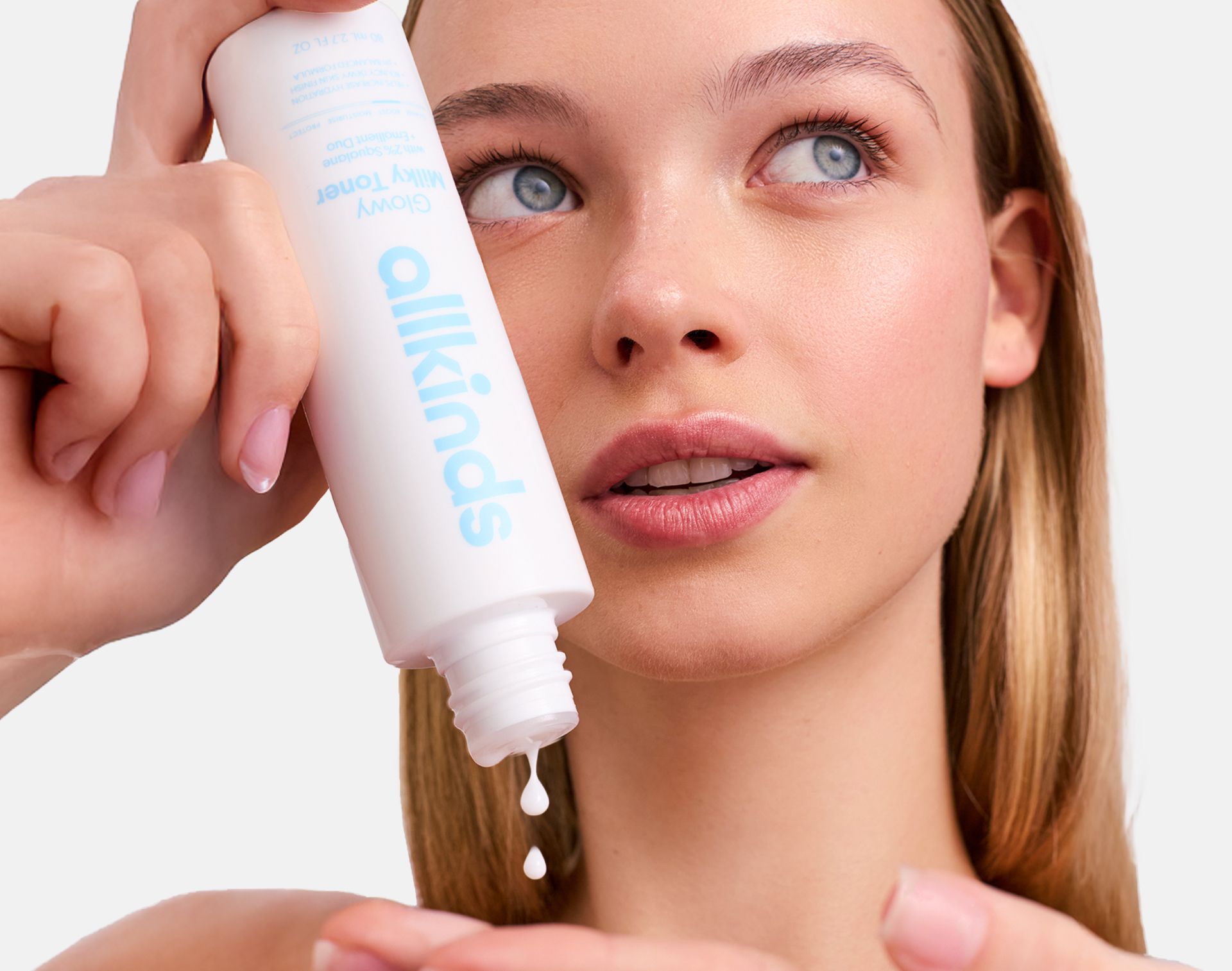 A woman pours "allkinds Glowy Milky Toner" from a white bottle into her hand.