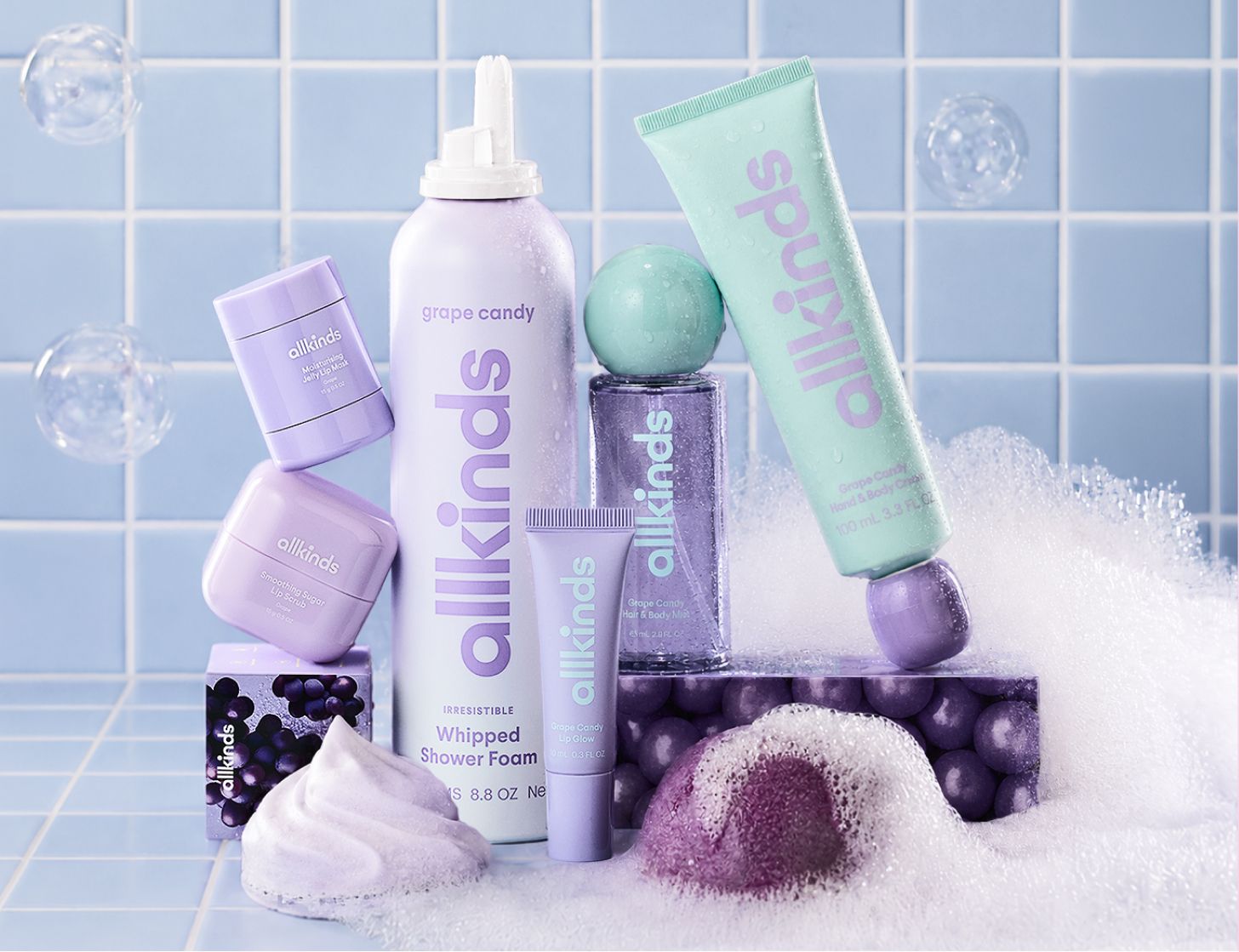 A collection of lavender and mint green allkinds bath products, including whipped shower foam and body wash, surrounded by bubbles on a blue tiled background.