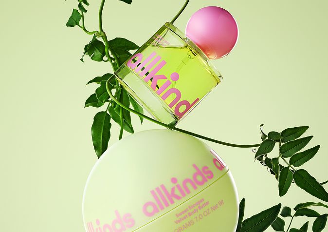 Allkinds perfume bottle and spherical body butter in green and pink, balanced amongst green leaves on a light green background.
