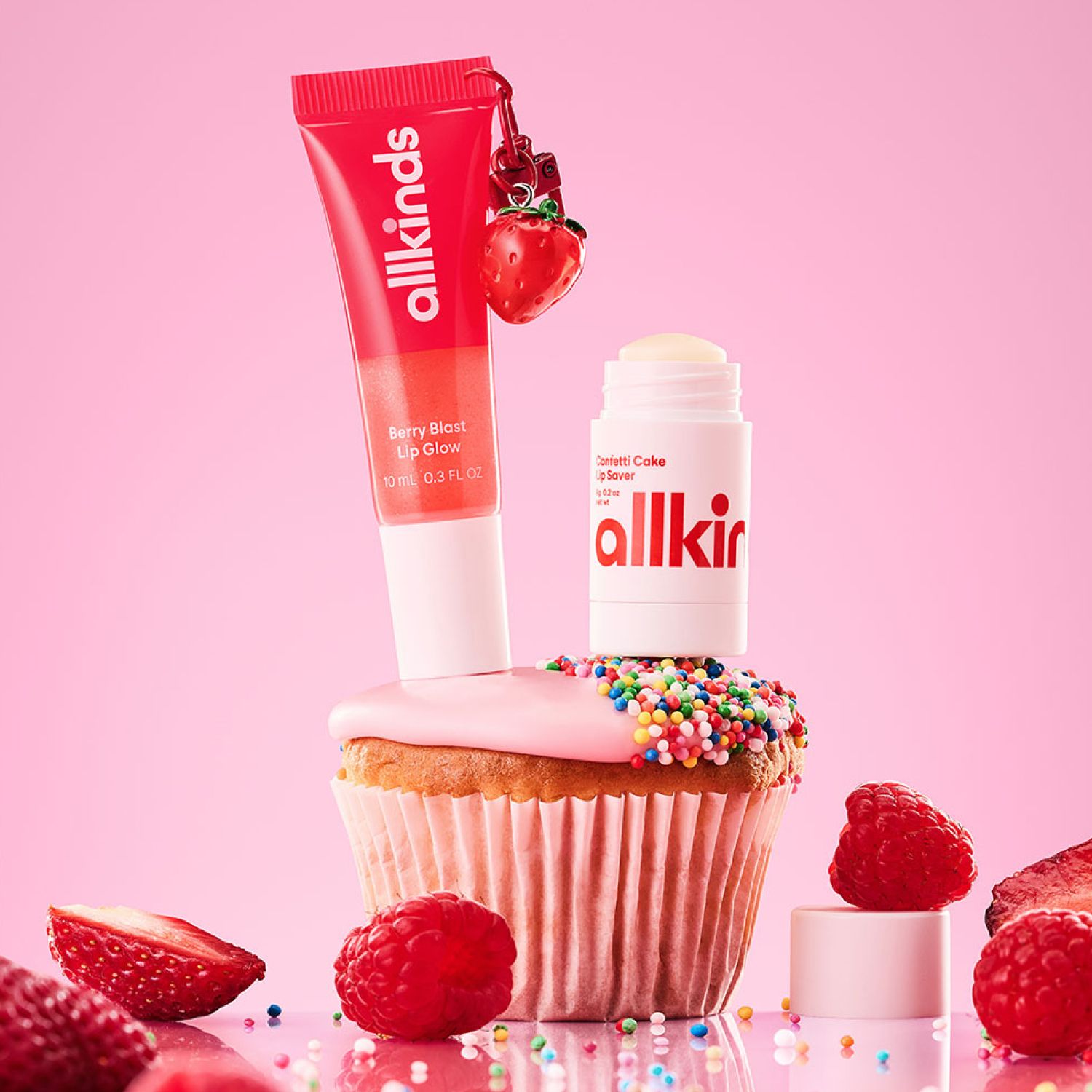 Allkinds Berry Blast Lip Glow and Confetti Cake Lip Saver balanced on a sprinkle-covered cupcake, surrounded by fresh berries on a pink surface.