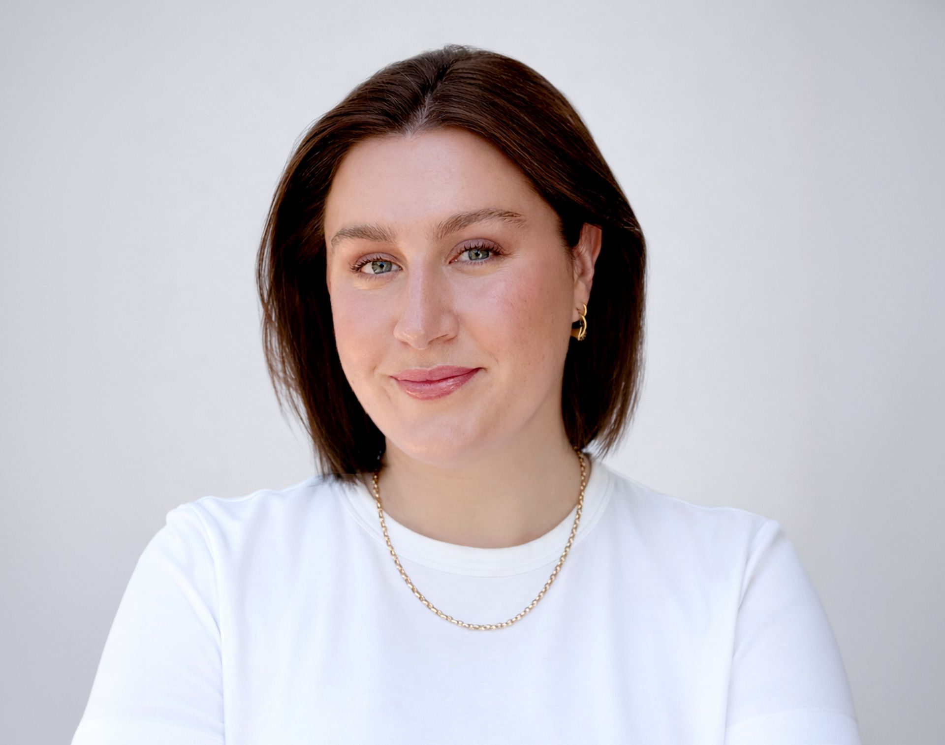 A young woman with short brown hair, a white t-shirt, and a gold necklace smiles softly.