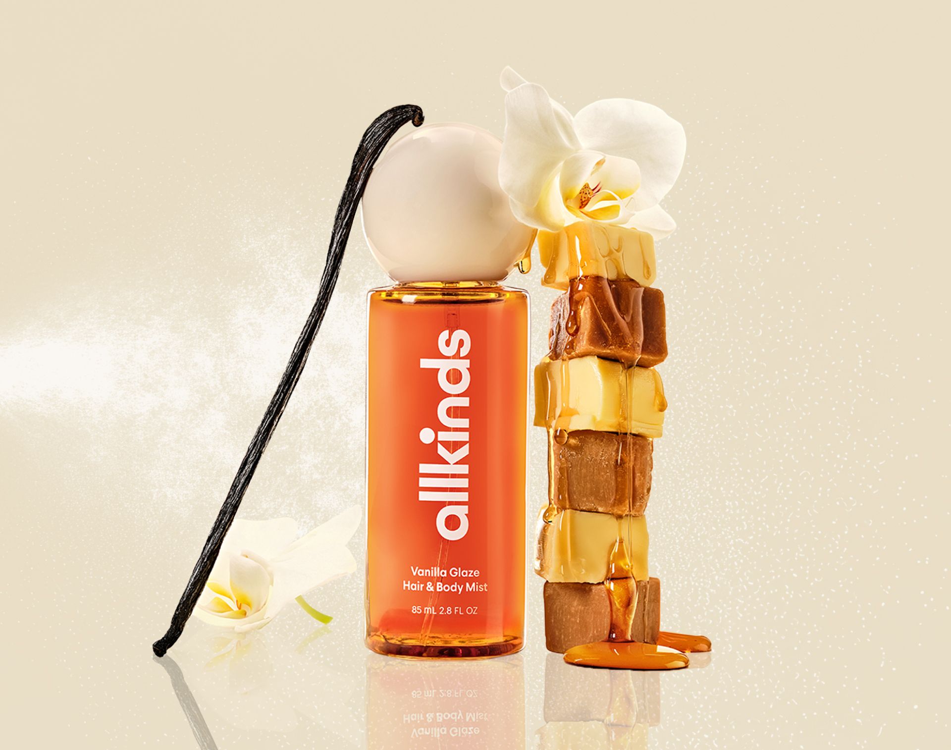 A bottle of allkinds Vanilla Glaze Hair & Body Mist with a vanilla bean, an orchid, and a stack of caramel squares drizzled with syrup.