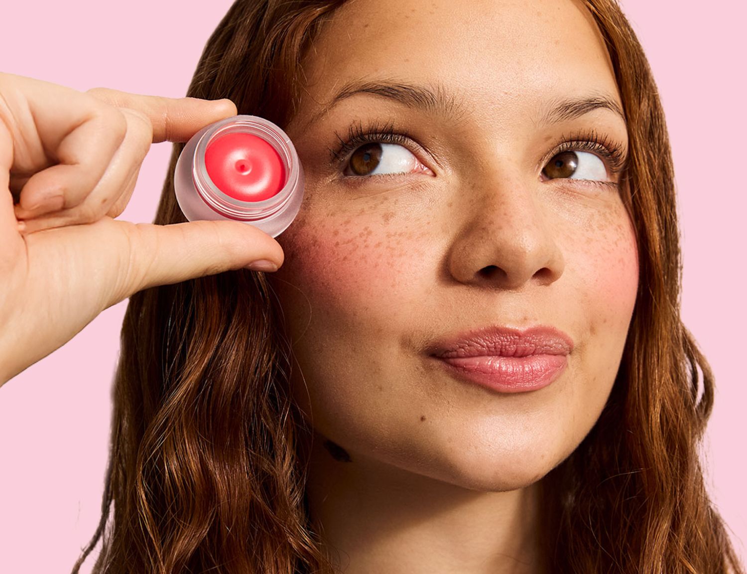 A woman with freckles and rosy cheeks looks up as a hand holds a small pot of pink blush near her temple.