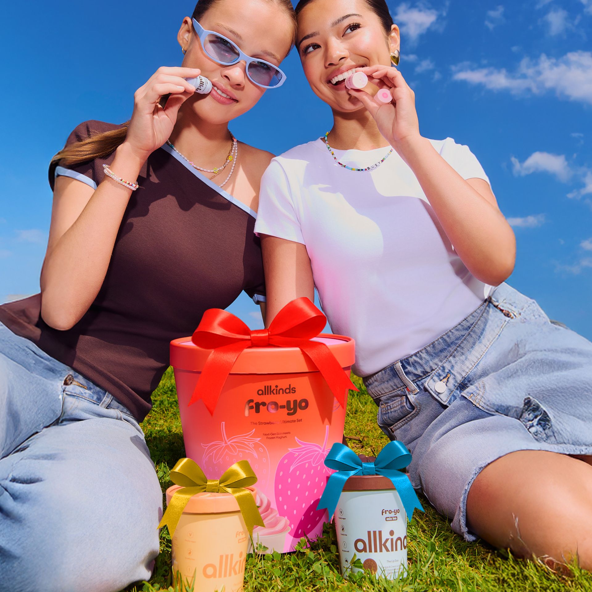 Two smiling women sit on grass, holding small round objects, beside three allkinds fro-yo containers with decorative bows.