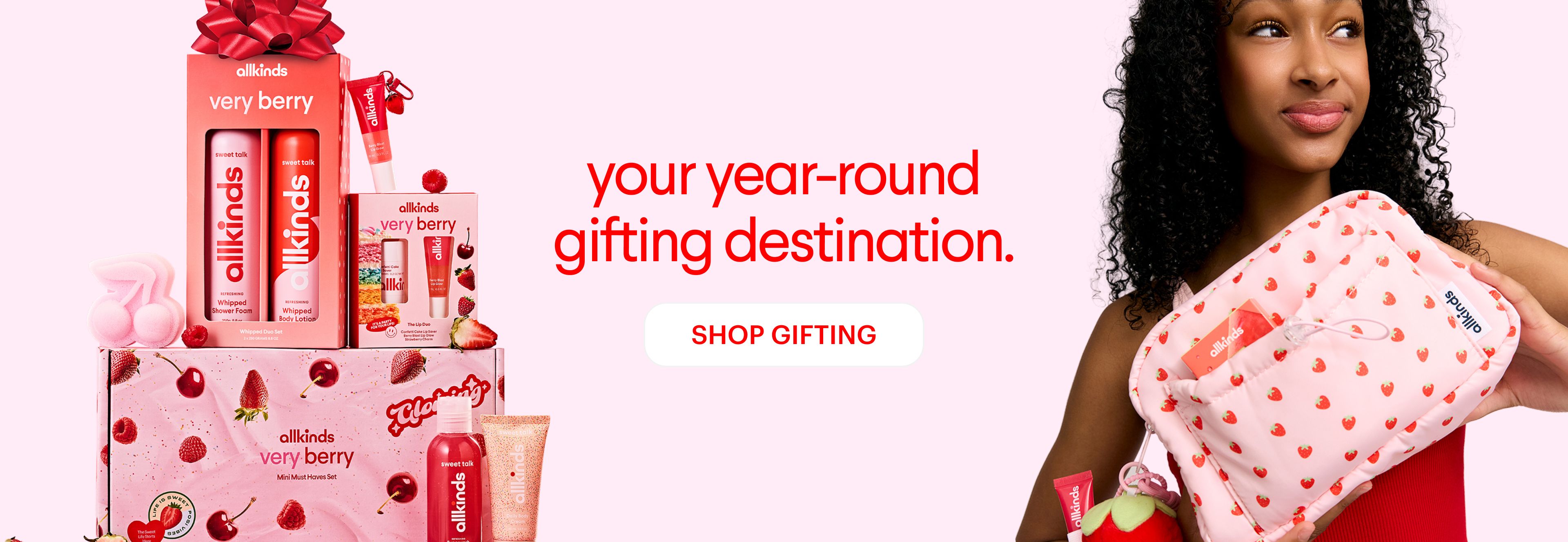A smiling woman holds a heart-patterned makeup bag next to a stack of berry-themed gift sets, with text "your year-round gifting destination. SHOP GIFTING."