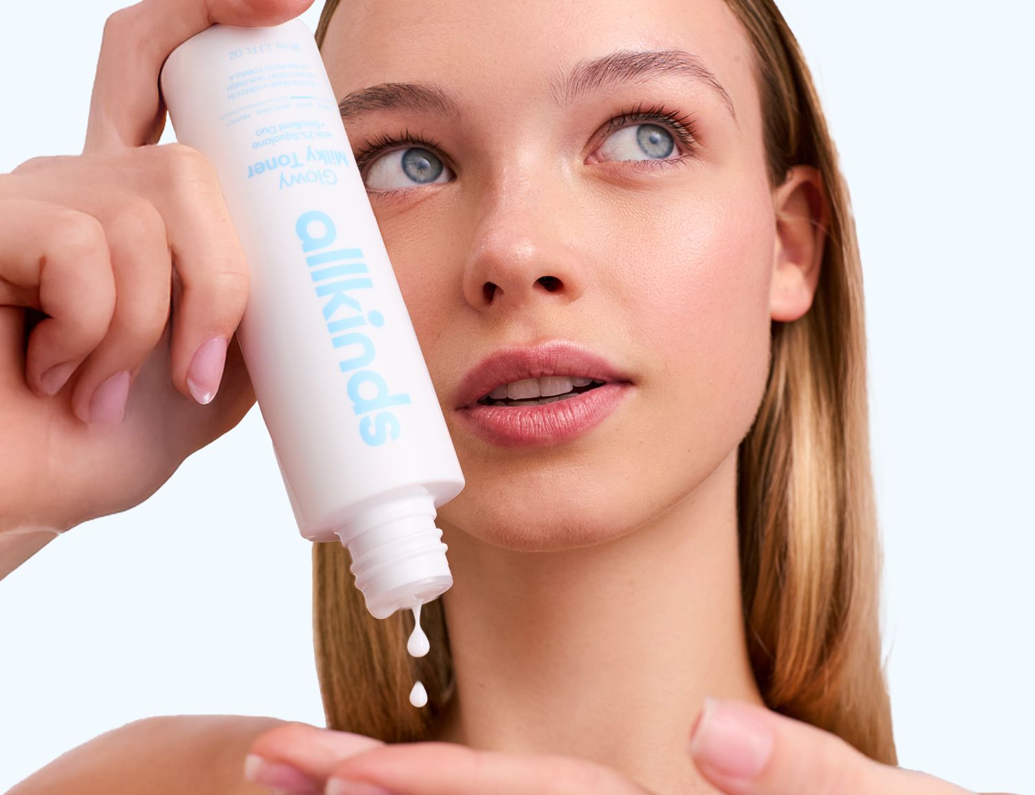 A young woman with blonde hair pouring "allkinds Glowy Milky Toner" from a white bottle into her hand.