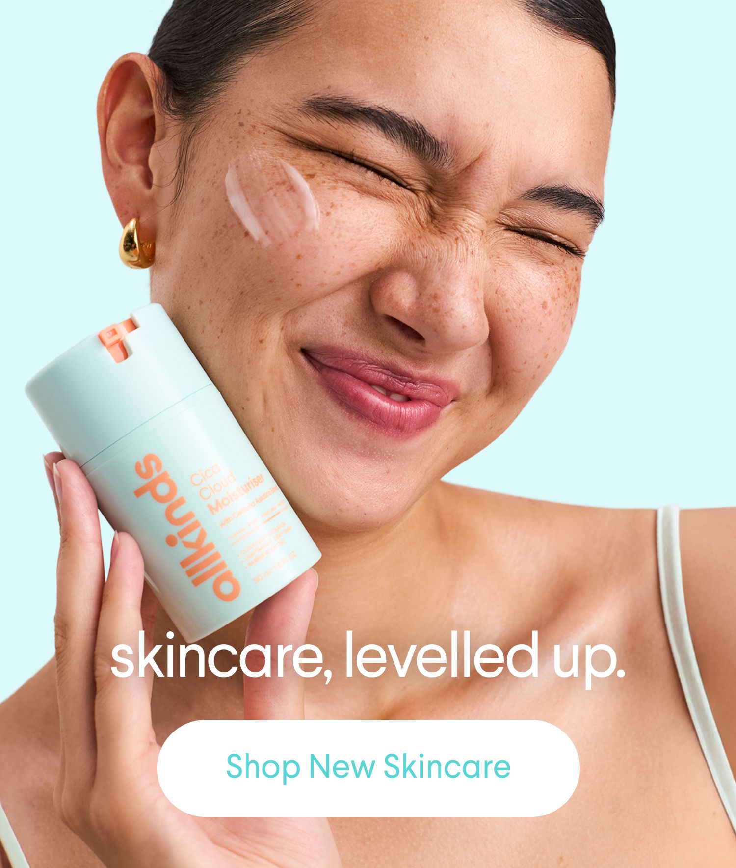 A joyful woman with freckles, eyes closed, has moisturizer on her cheek and holds an allkinds Cica Cloud Moisturiser bottle.