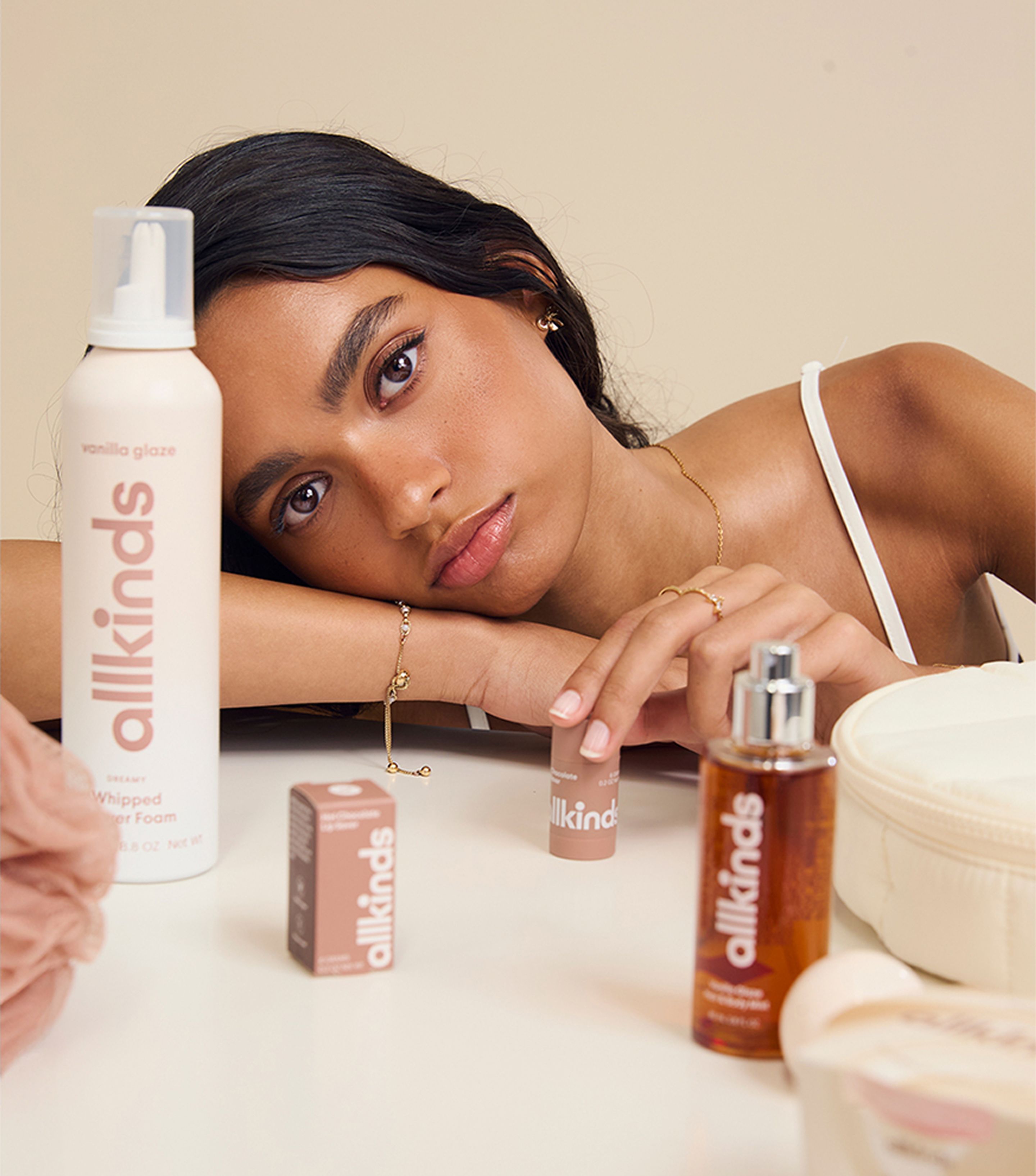 A woman with dark hair rests her head on her arms, looking at the camera, surrounded by "allkinds" brand body care products, including a shower foam, a small stick, and an oil spray.