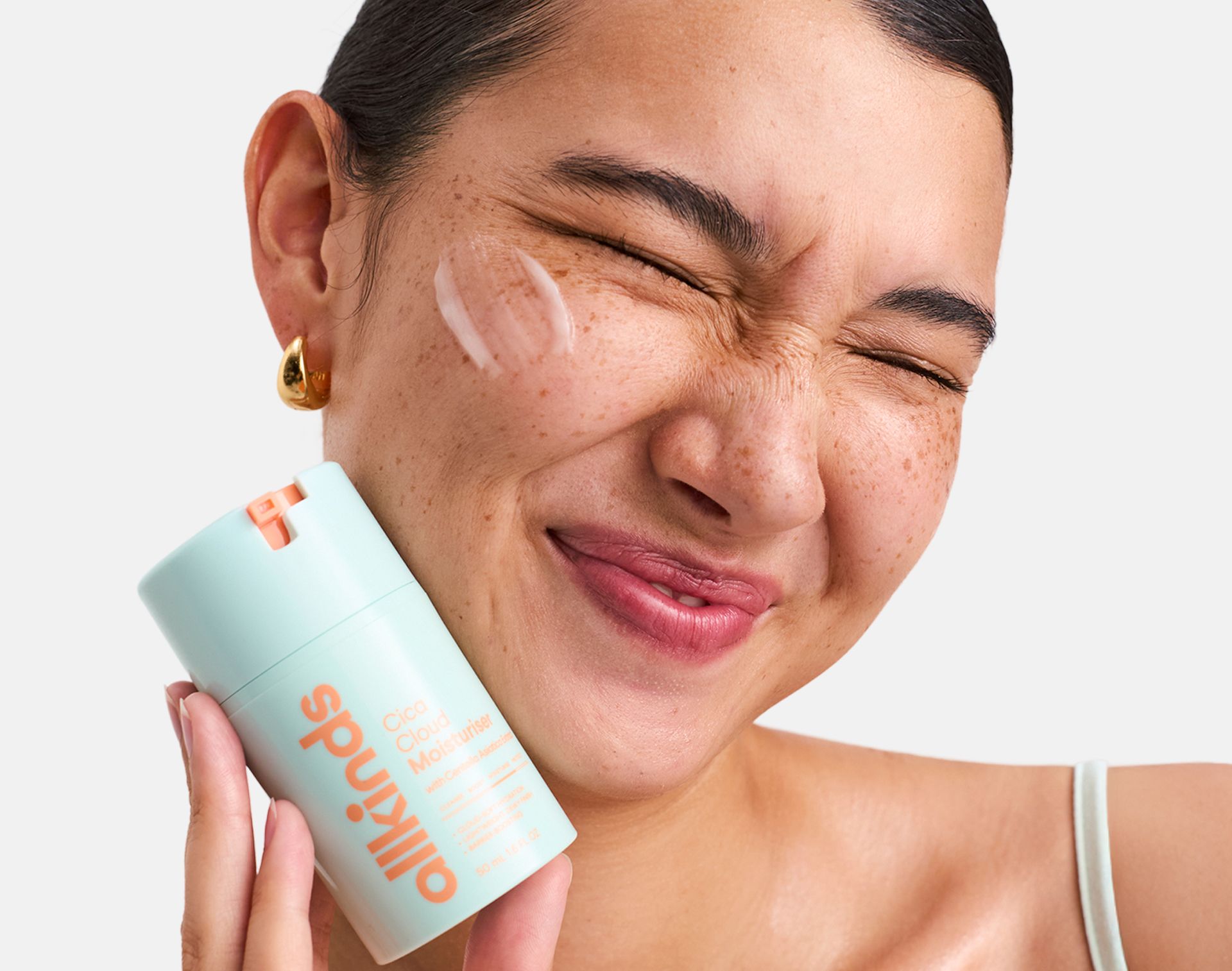 Joyful woman with freckles holding an Allkinds Cica Cloud Moisturizer, with cream on her cheek.