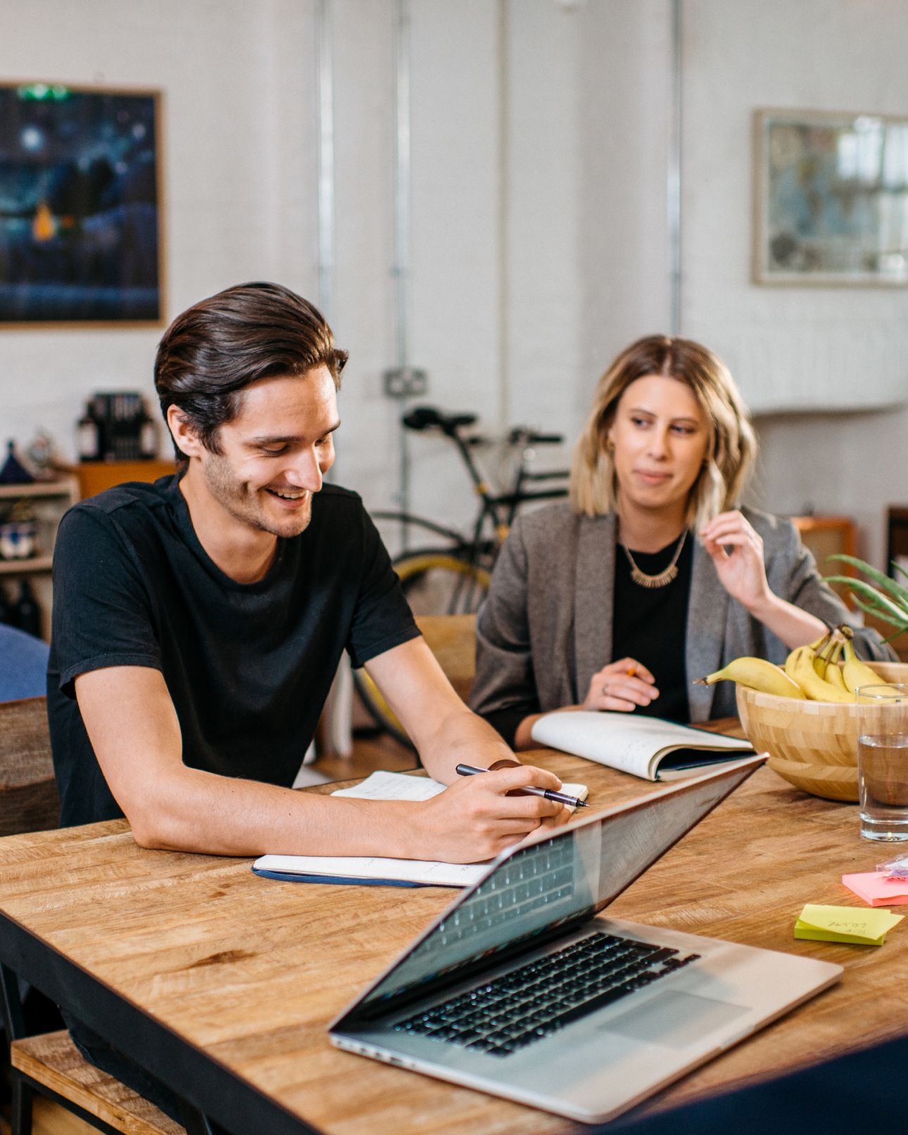 Two team members at a wooden table with a laptop and notebooks, one smiling while writing. AI & Emerging Technology.
