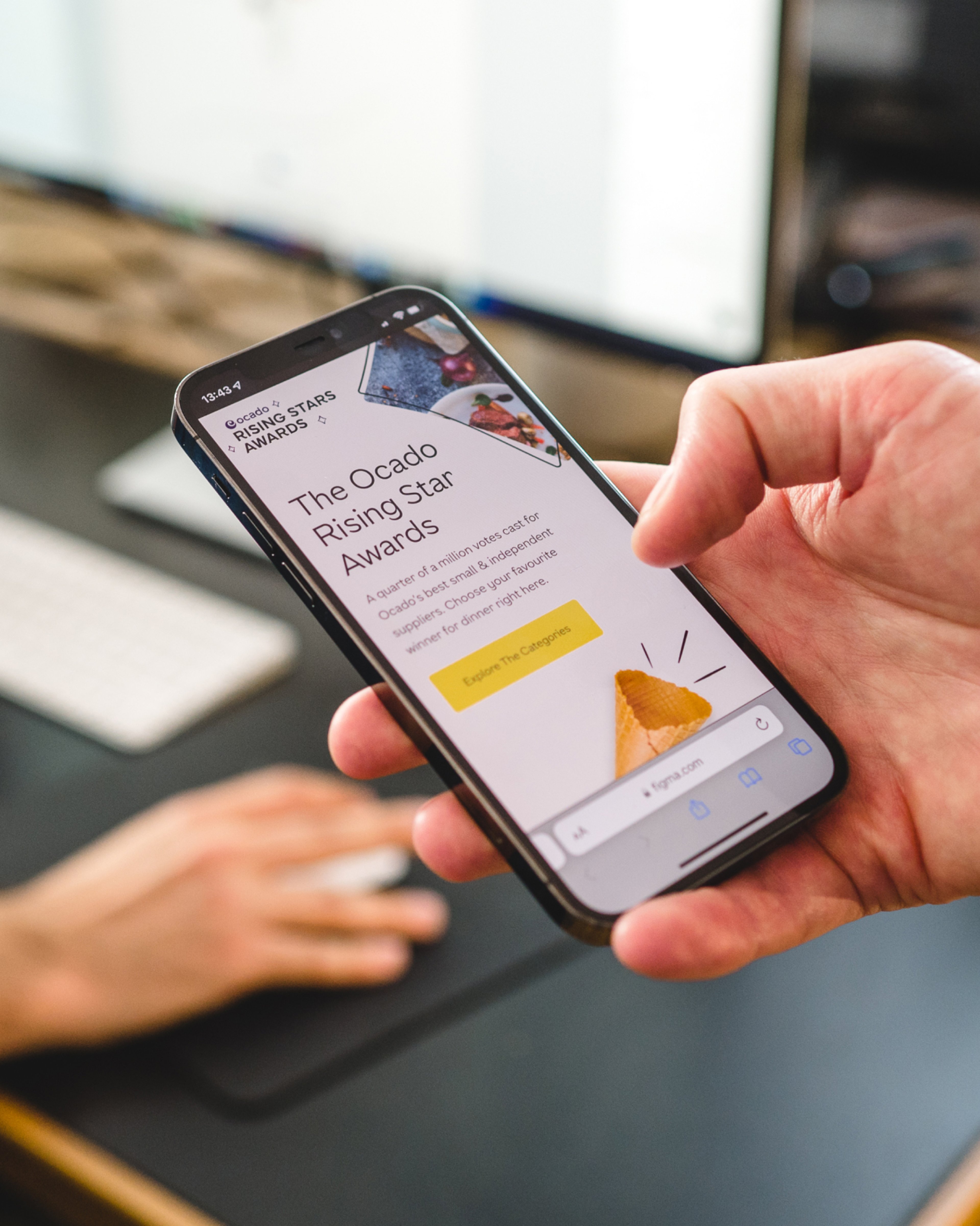 A hand holding a phone showing an awards website, with a desktop computer behind. Marketing and eCommerce websites, Software Engineering.
