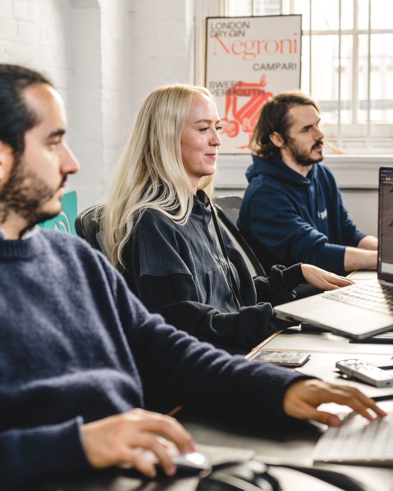 Team members working side by side at computers in the studio, a vintage poster on the wall behind them. Experience Design.