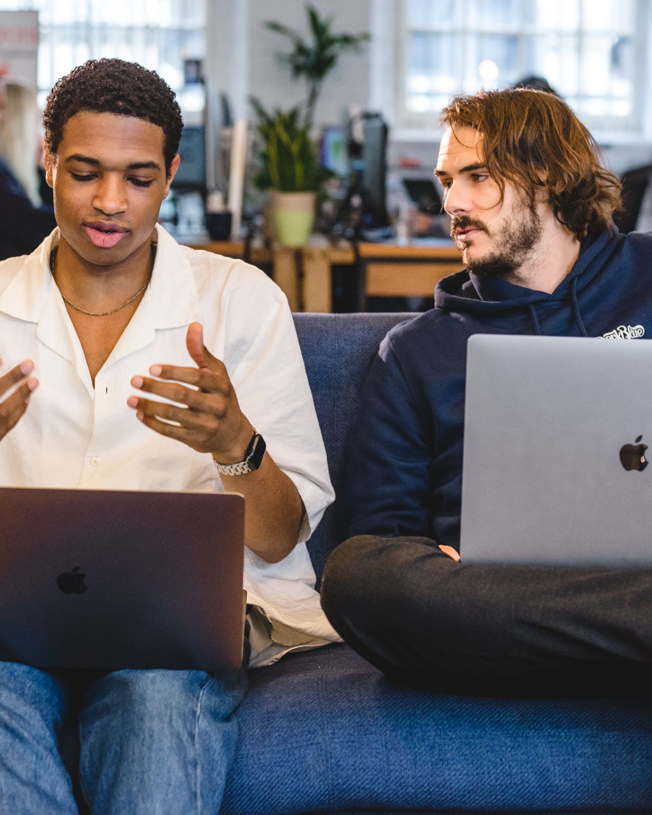 Two team members talking on a sofa with laptops, one gesturing as they explain. AI product development, AI & Emerging Technology.