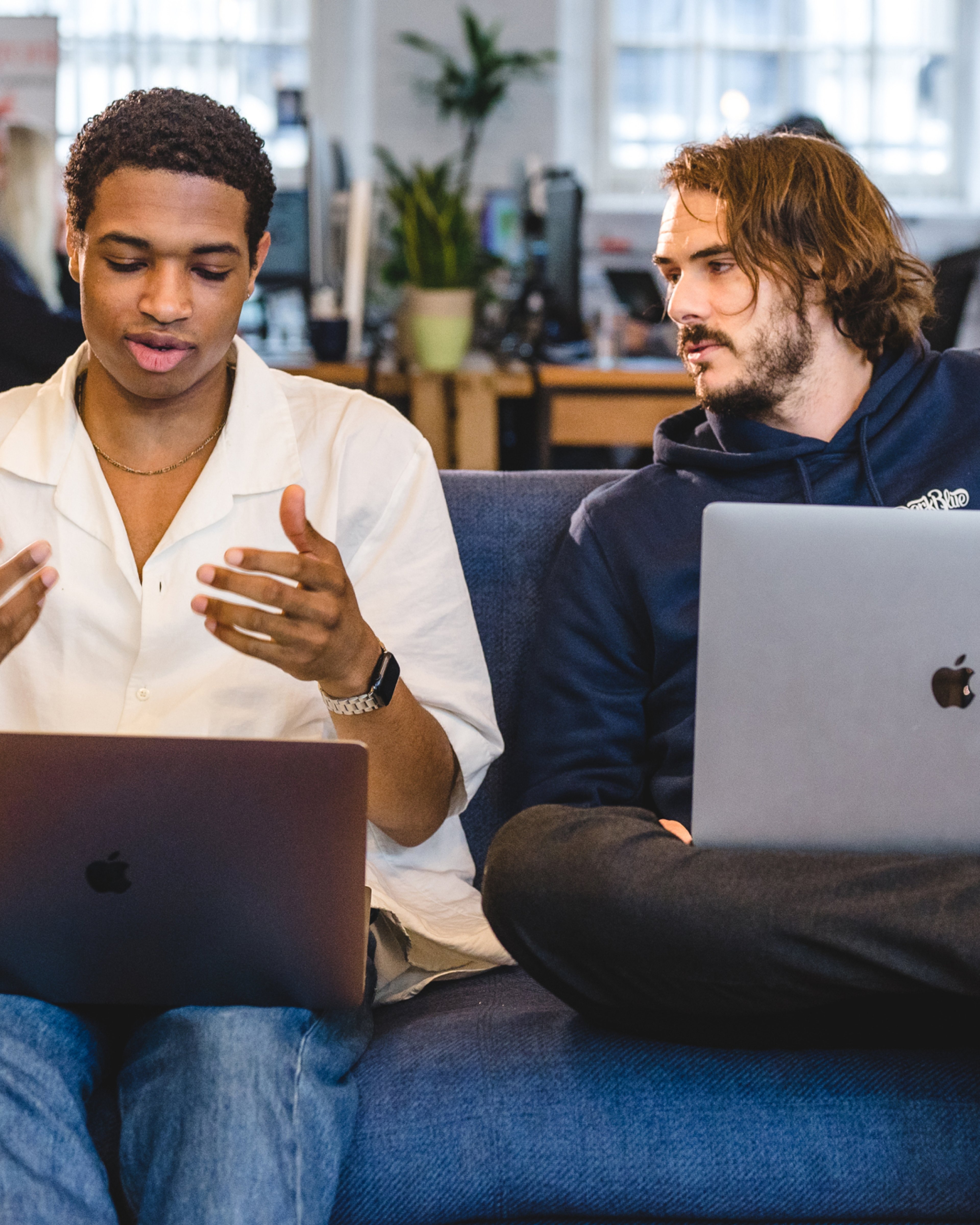 Two team members talking on a sofa with laptops, one gesturing as they explain. AI product development, AI & Emerging Technology.
