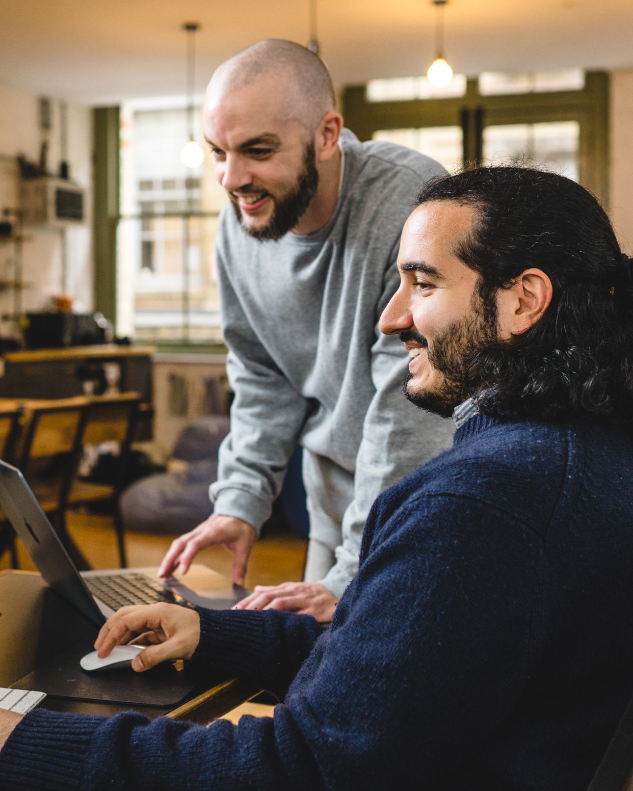 A team member leaning over a colleague's shoulder as they both smile at a laptop screen. Rapid ‘proof of concept’ testing, Research & Strategy.