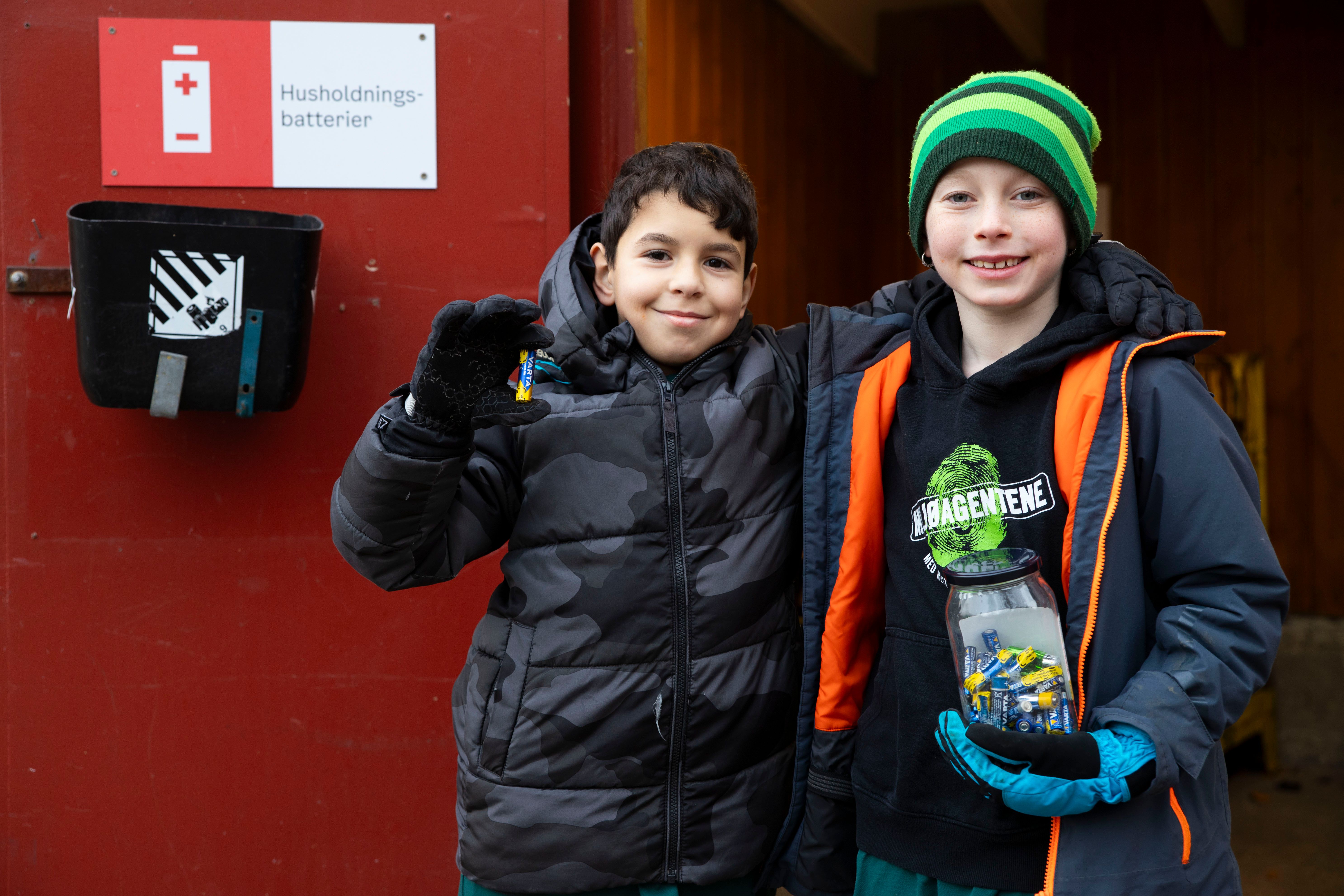 To gutter i vinterklær står utenfor en gjenvinningsstasjon. Gutten til høyre holder et syltetøyglass med brukte batterier, gutten til venstre holder et batteri opp mot kamera. Til høyre for guttene er en rød dør hvor det henger en svart boks og et skilt med sorteringsmerket for batterier og teksten husholdningsbatterier
