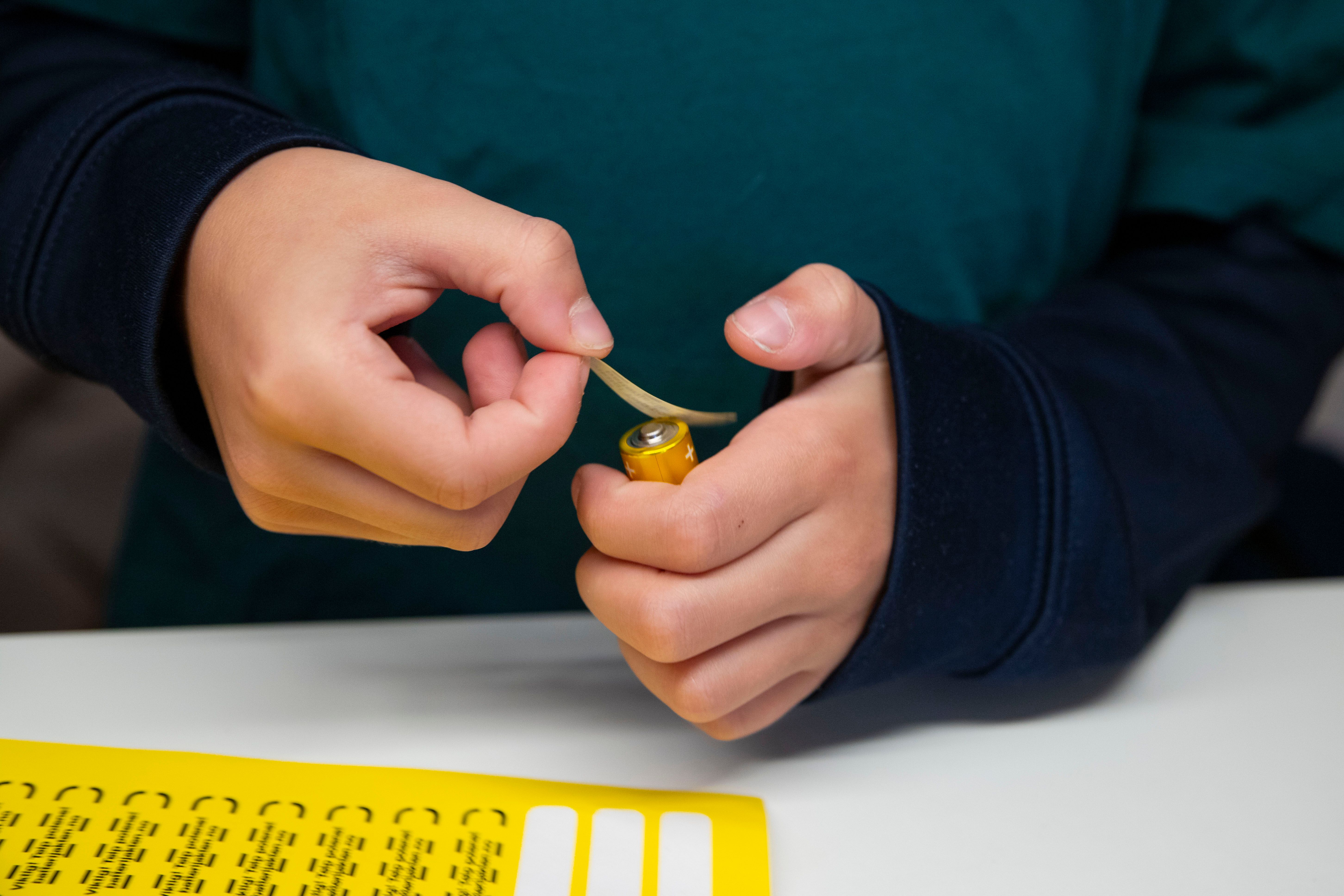 Nærbilde av hender. I den ene hånda er det et AA-batteri, og den andre hånda holder en teipbit som er iferd med å teipes over batteriet. I forgrunnen er det gule teipe-klistremerker