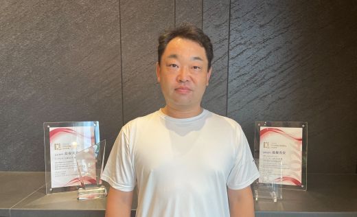 賞状とトロフィーの前に立ち緊張気味の男性の写真