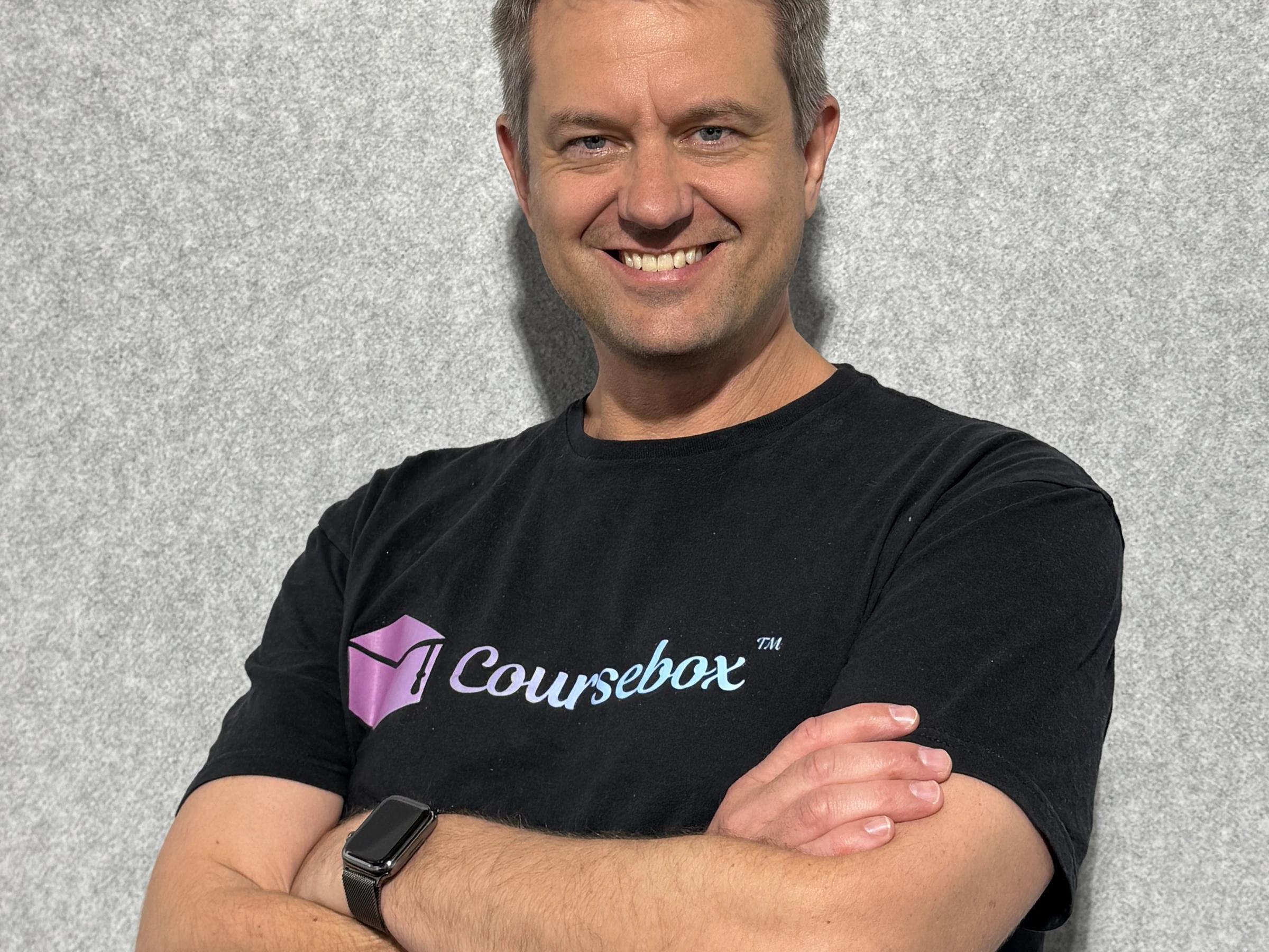 Travis Clapp in a black Coursebox T-shirt, arms crossed, smiling in front of a grey wall