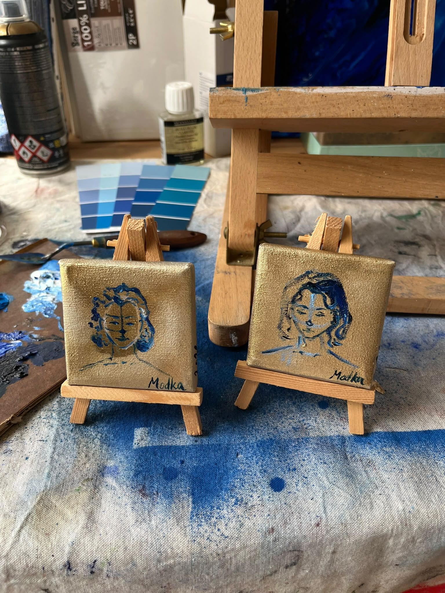 Deux toiles miniatures à fond doré sur de petits chevalets, dans l’atelier, devant un nuancier de bleus.