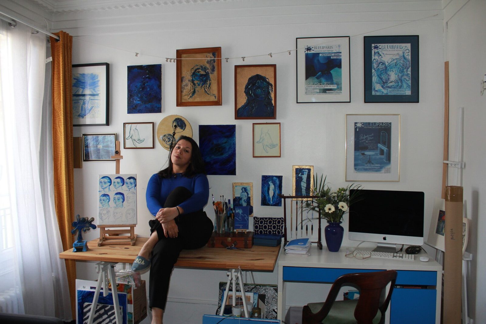 Modka assise sur sa table de travail, dans son atelier parisien, devant un mur couvert de ses œuvres bleues et des affiches de Bleu Paris.