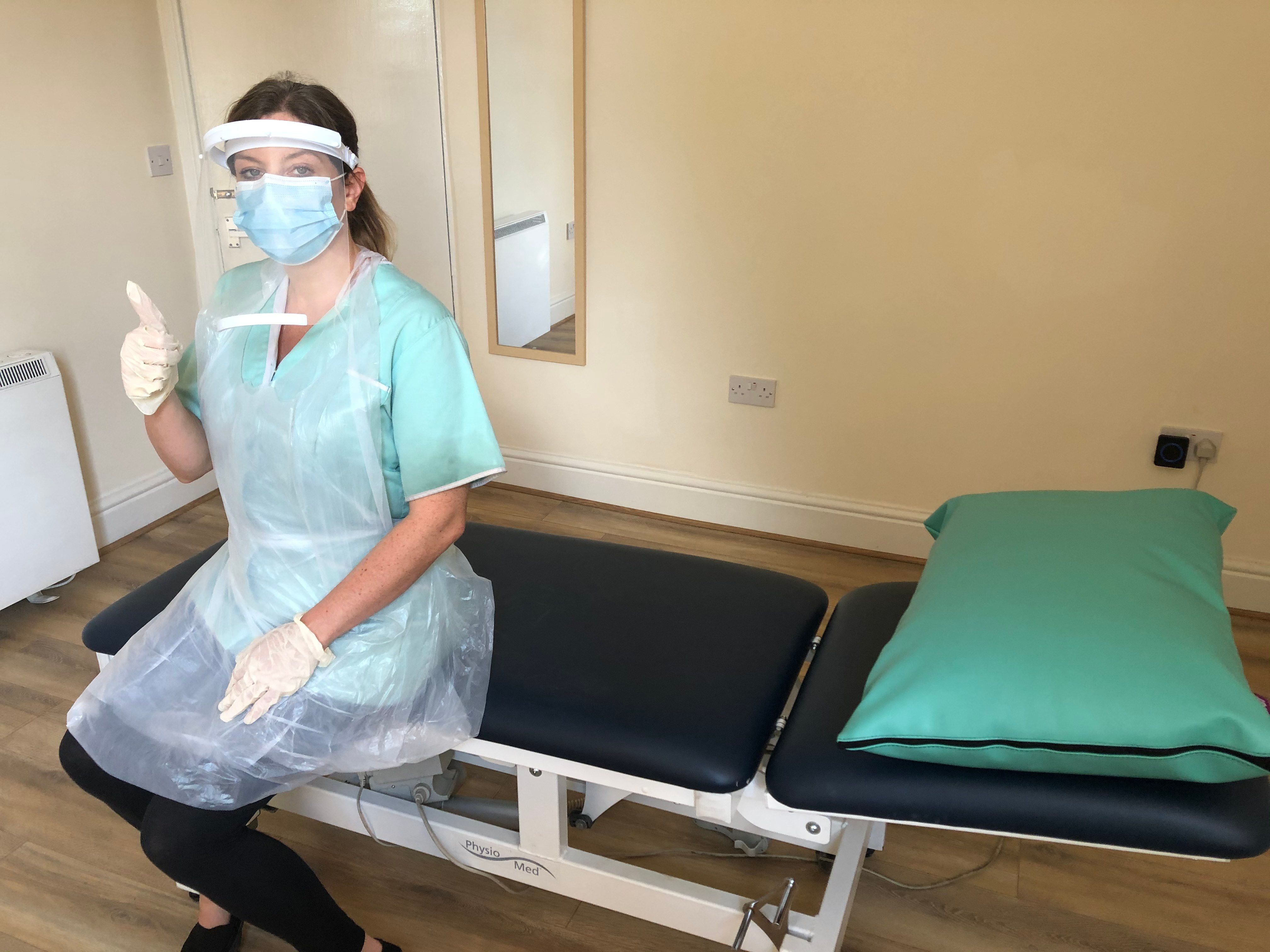 Collette Bourke (B.OST) sitting on her treatment table wearing clinic clothes, a plastic apron, nitrile gloves, a visor and mask. 