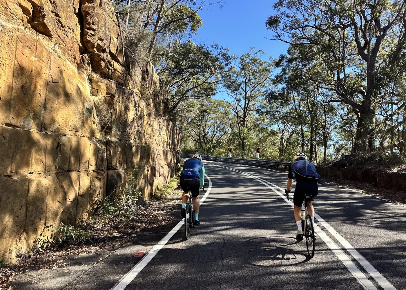 Two riders on a quiet road cut through gold sandstone, gum trees overhead