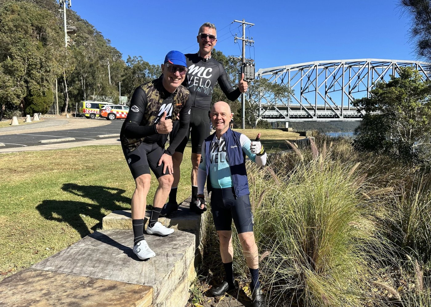 Three MC Velo riders posing on a ledge in front of the Hawkesbury bridge at Brooklyn
