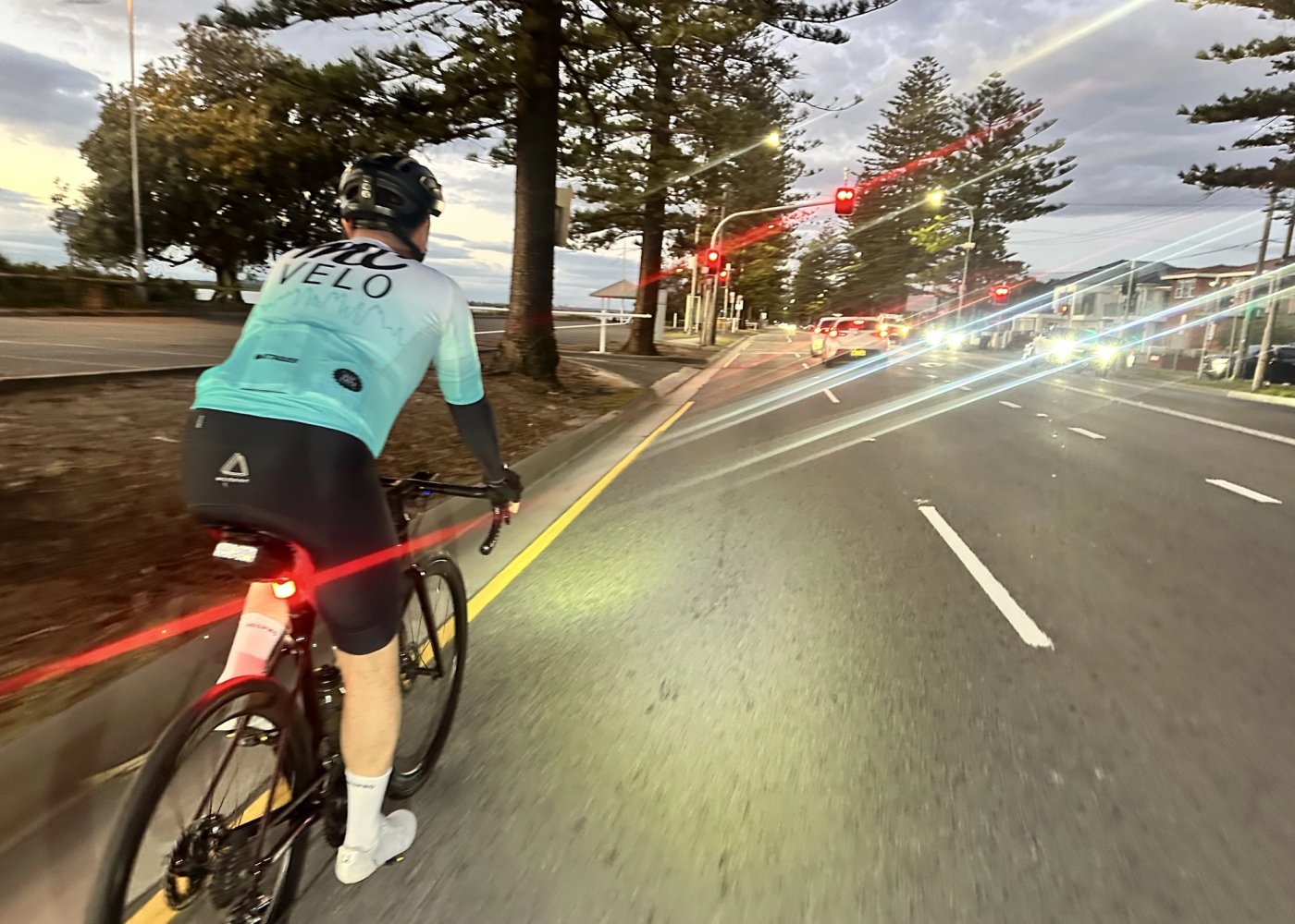 A rider in MC Velo kit heading south along the coast road at dawn, rear light streaking, Norfolk pines and traffic lights ahead.