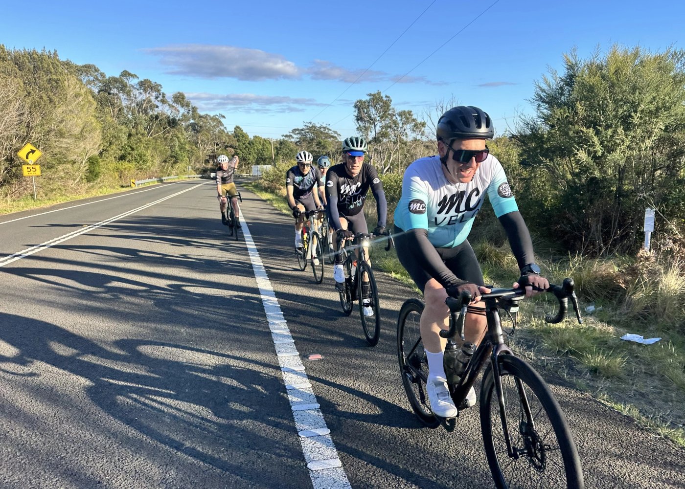 A line of riders on an open road in the sun, the group strung out behind the rider on the front.