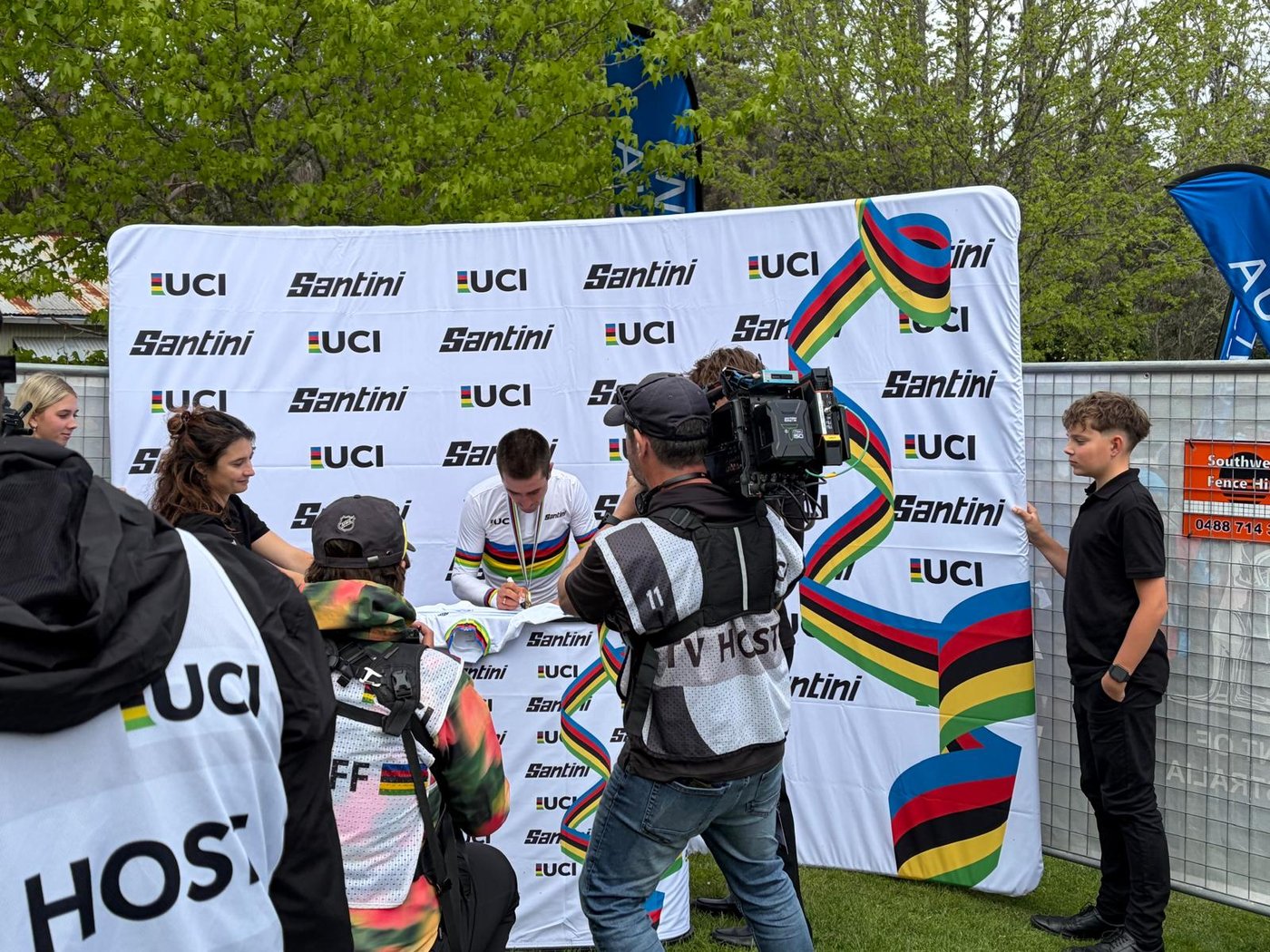 Magnus Bak Klaris in the rainbow jersey signing a jersey at the UCI backdrop, a TV camera alongside