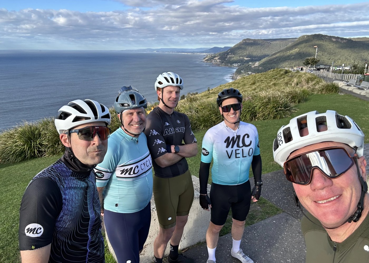 Five riders in MC and MC Velo kit at a clifftop lookout, the coastline and escarpment stretching north behind them.