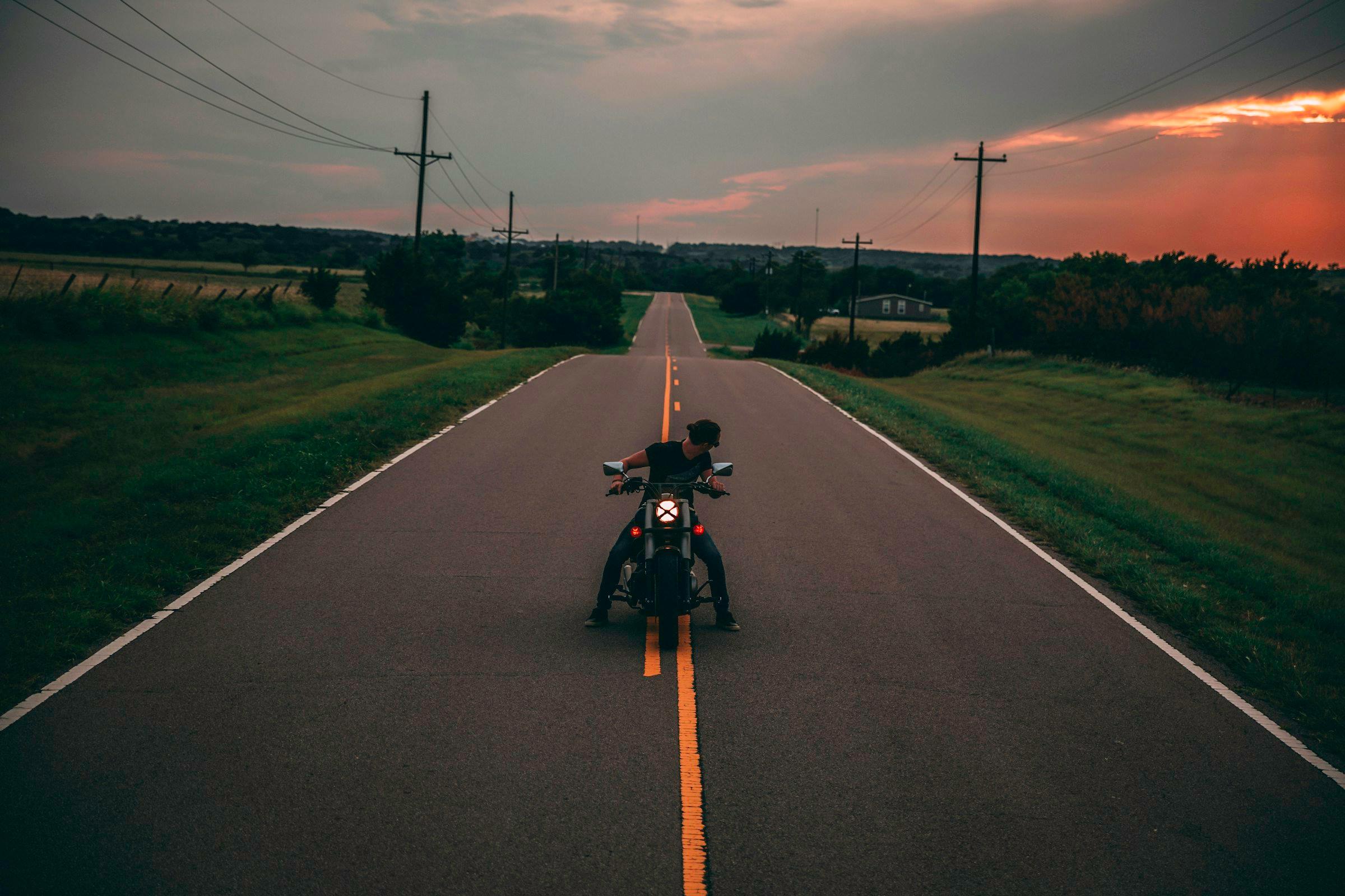 Motorcyclist riding down an empty two-lane rural road at sunset, with power lines and fields on either side